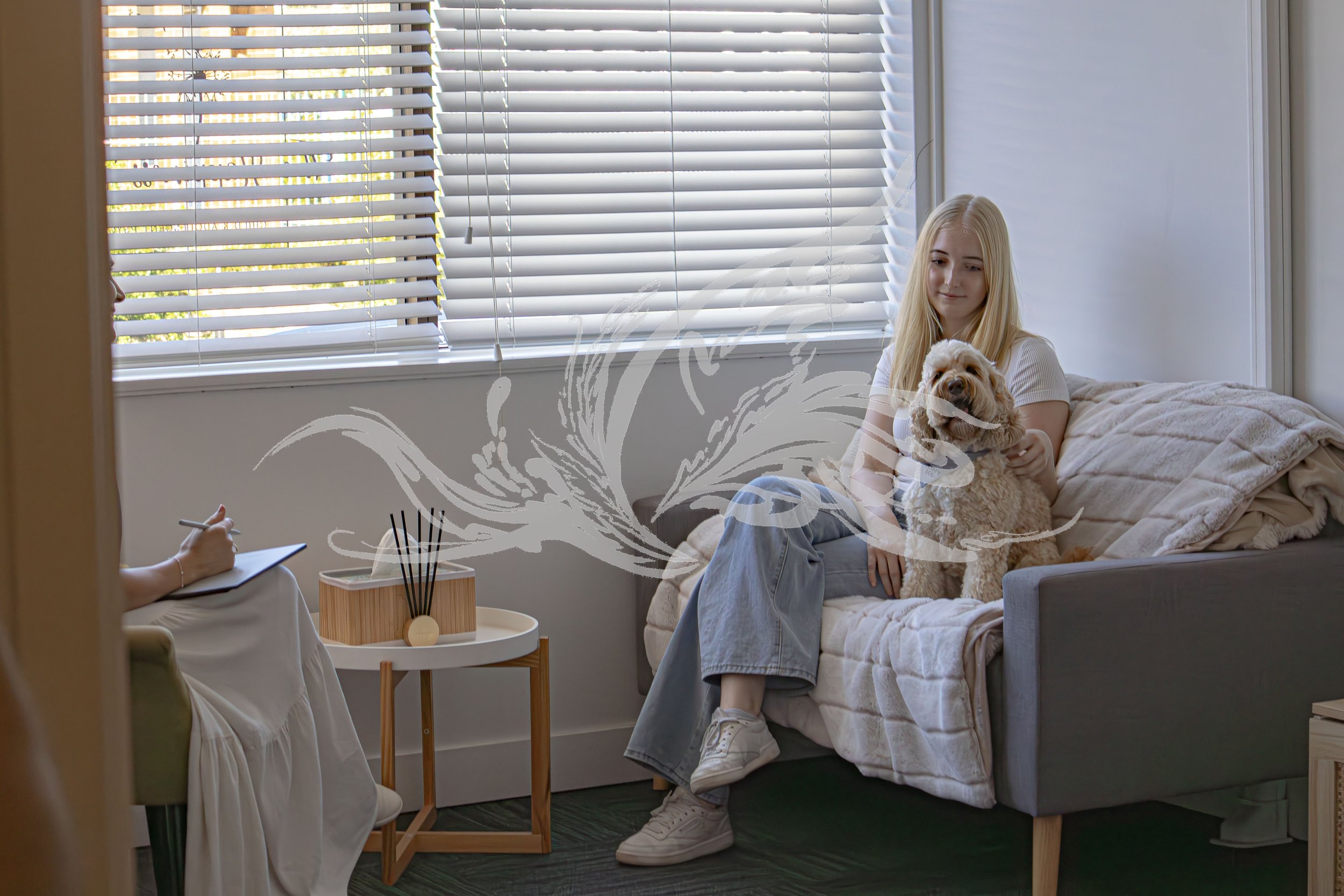 A young woman with long blonde hair sitting on a bed with a dog in her lap, in a bedroom with large window blinds and sunlight coming through.