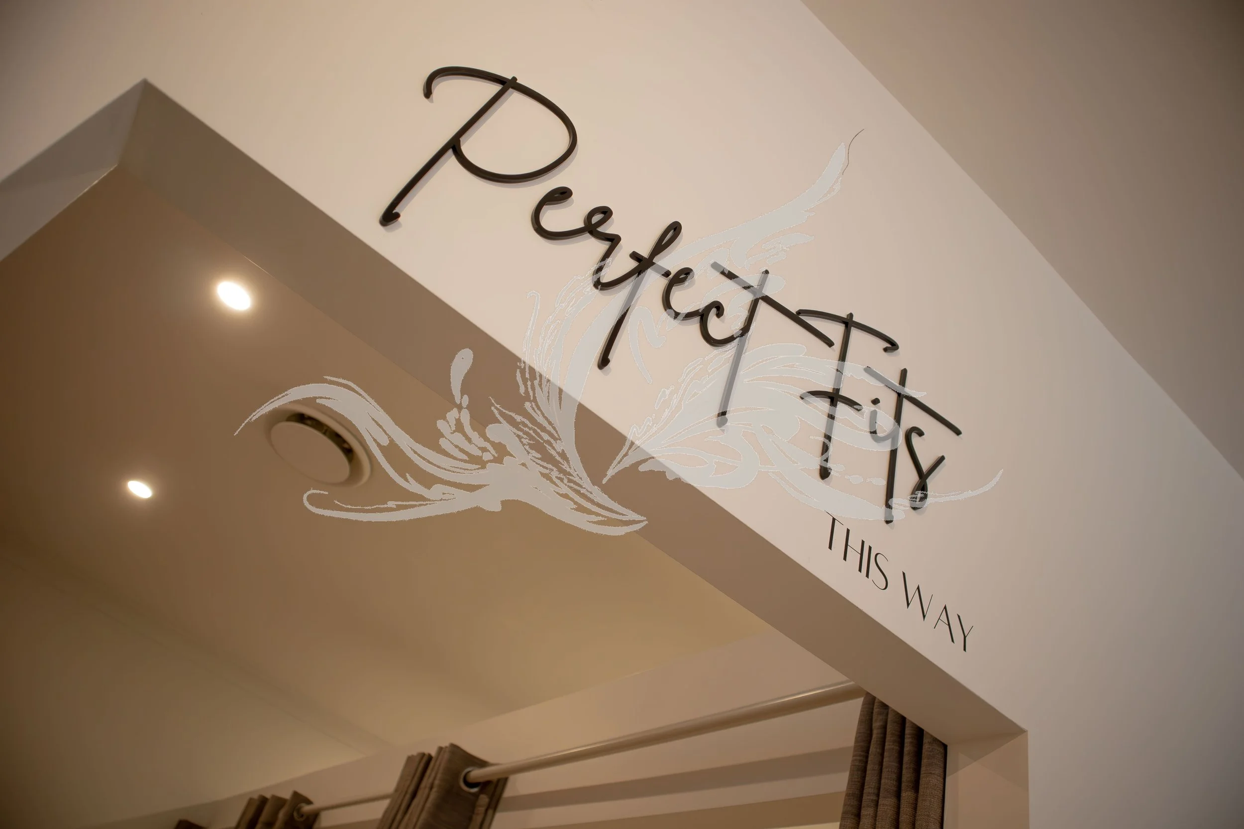 Close-up of a wall sign with cursive letters that read 'Perfect Fit, this way' and a decorative leaf design underneath.