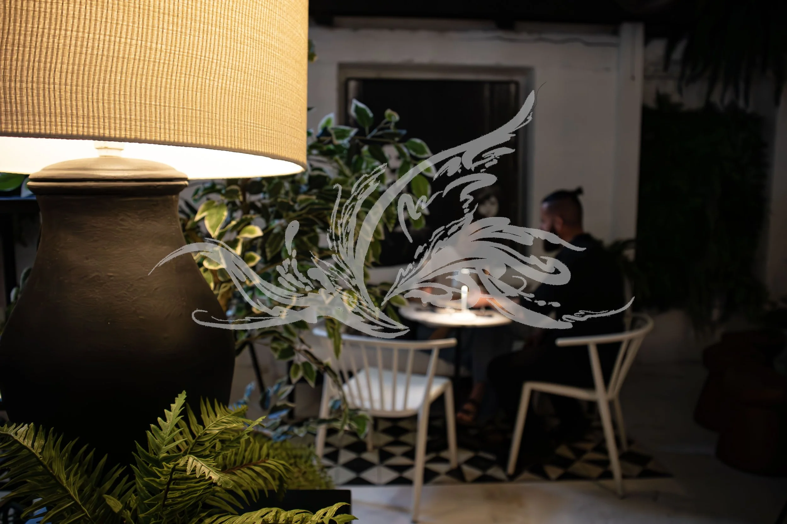 Indoor scene with a beige lampshade on a dark brown ceramic lamp base, green and variegated plants in the foreground, and a person with dark hair sitting at a table in the background. The space has black and white patterned flooring and white chairs.