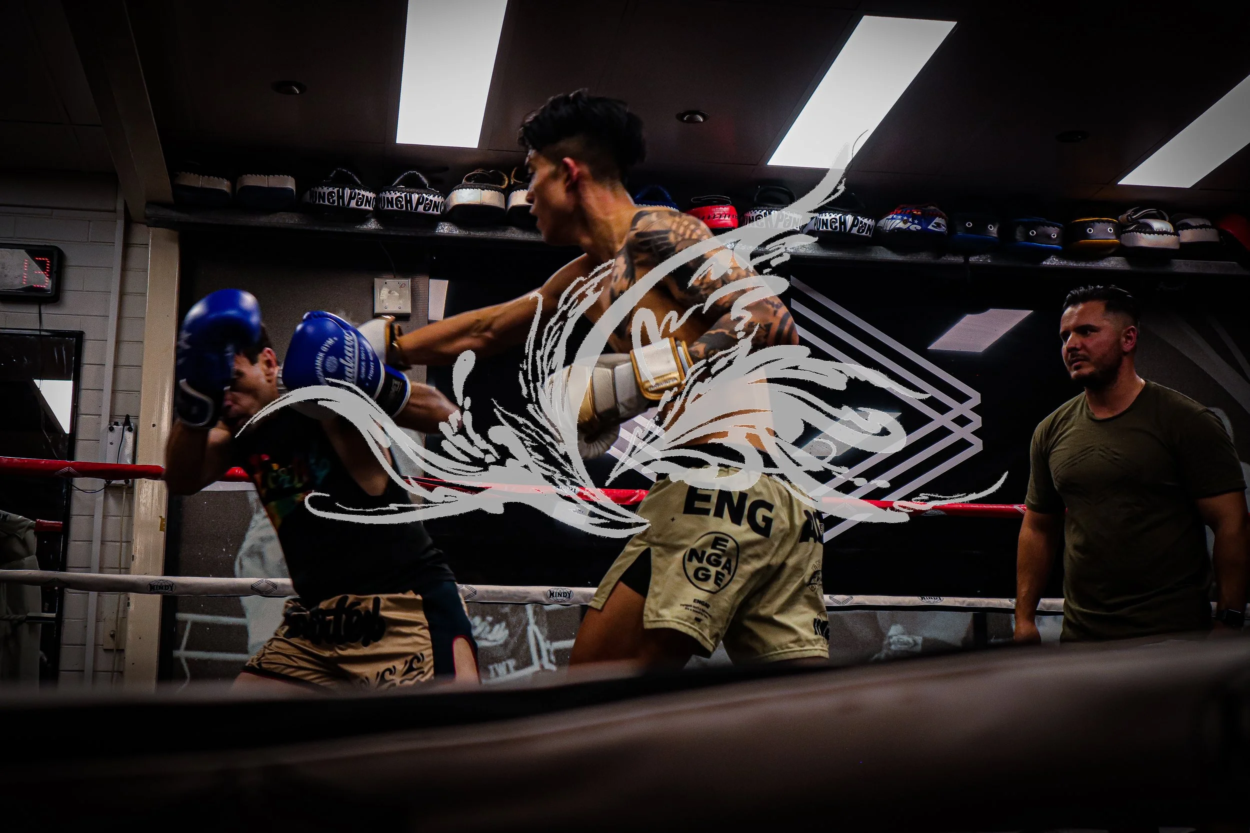 Two men practicing boxing in a gym with gloves, one delivering a punch while the other blocks. There is a person observing and a shelf above with boxing gloves.
