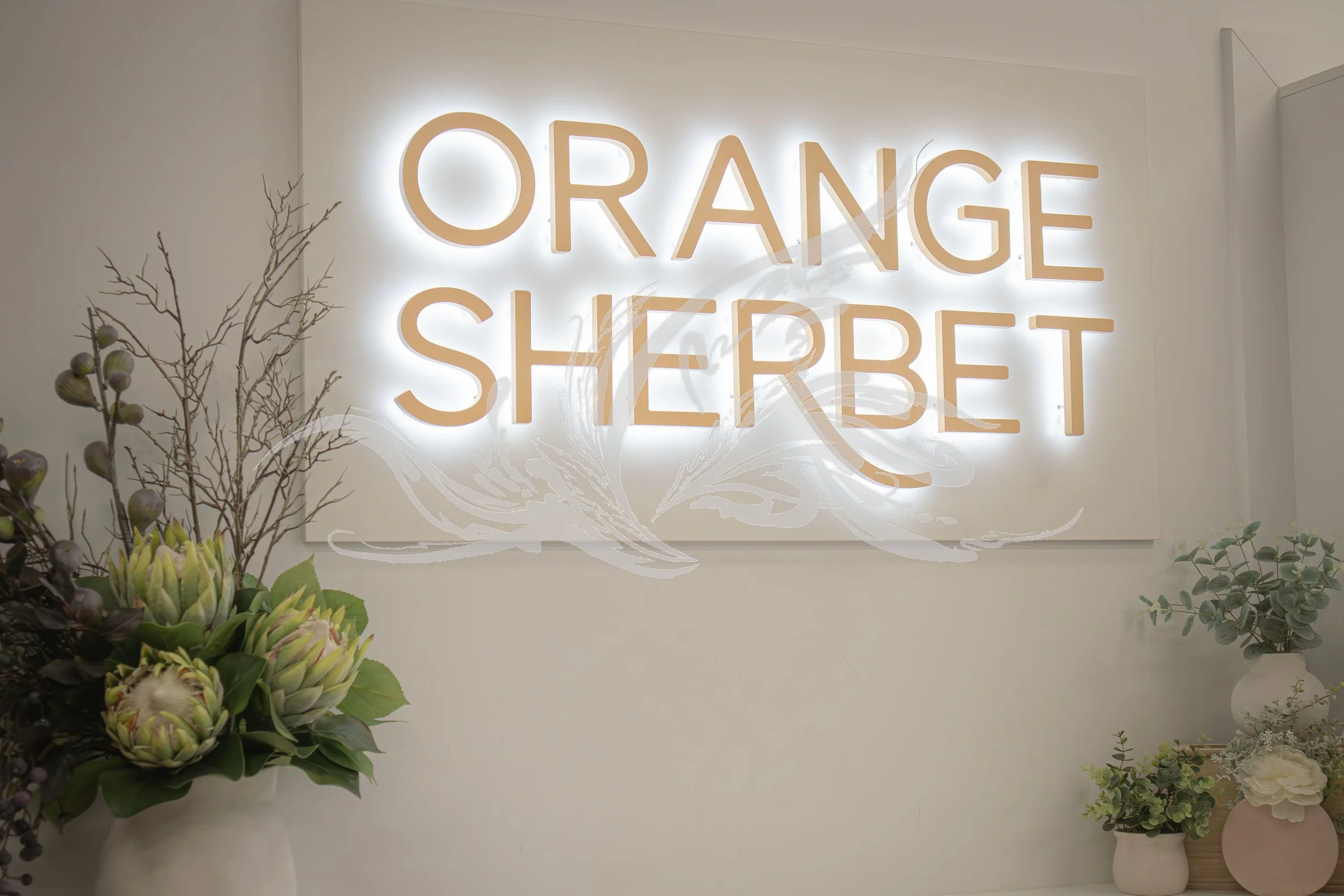 Decorative floral arrangement with a neon sign reading 'ORANGE SHERBET' in the background, along with plant vases and branches.