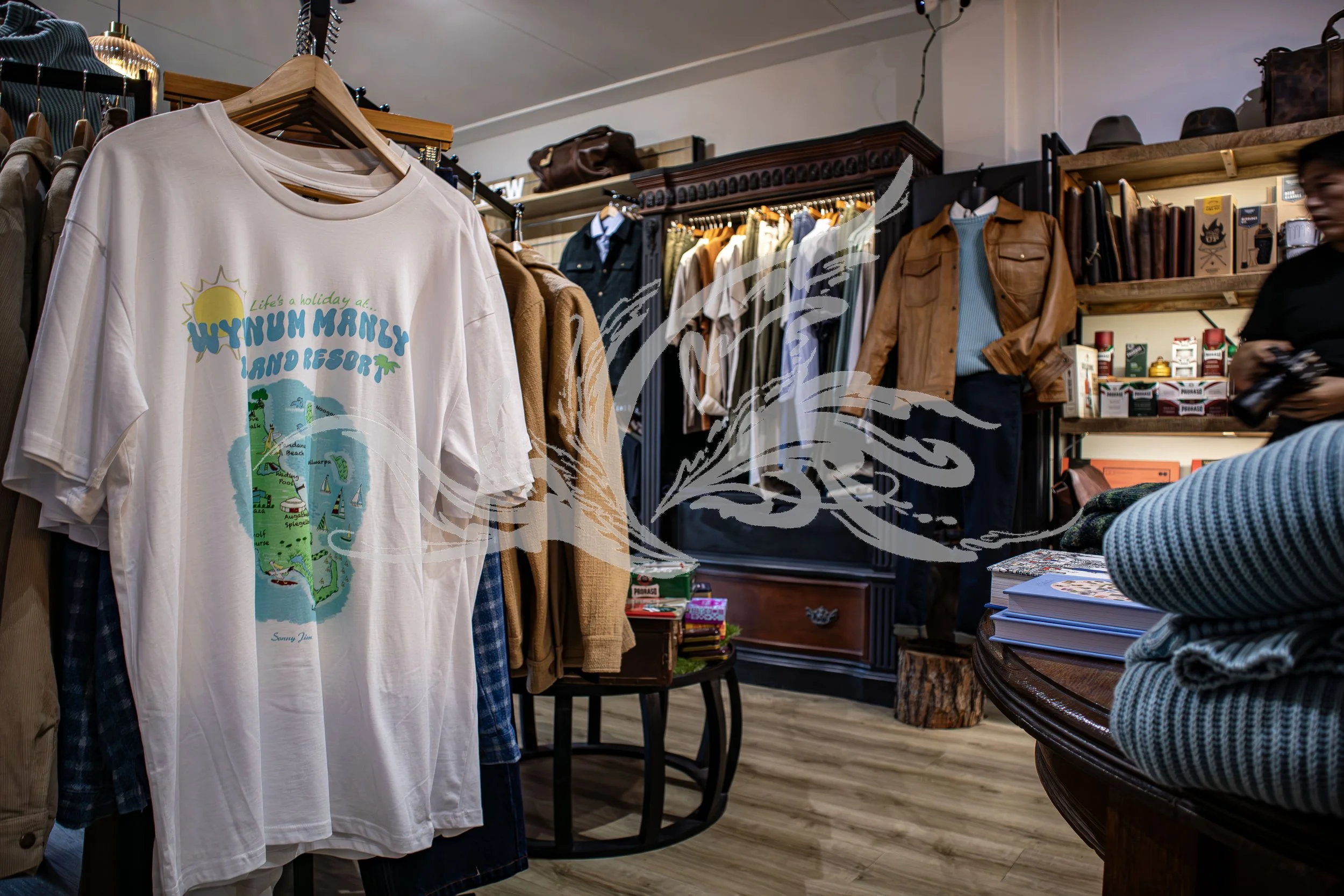 A clothing store display with a white t-shirt featuring a colorful summer-themed graphic and text, hanging on a wooden hanger among other garments.