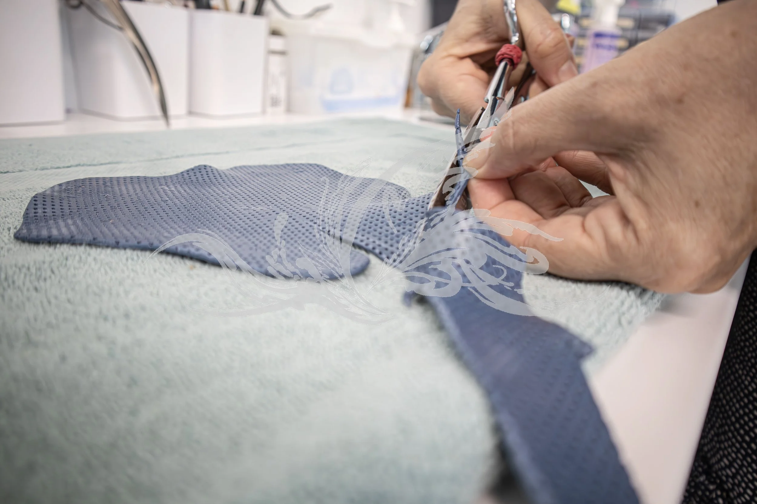 A person with visible veins on their hands is using a surgical scissors to cut a small pink surgical thread while wearing a latex glove. An blue textured pad is placed on the surface.