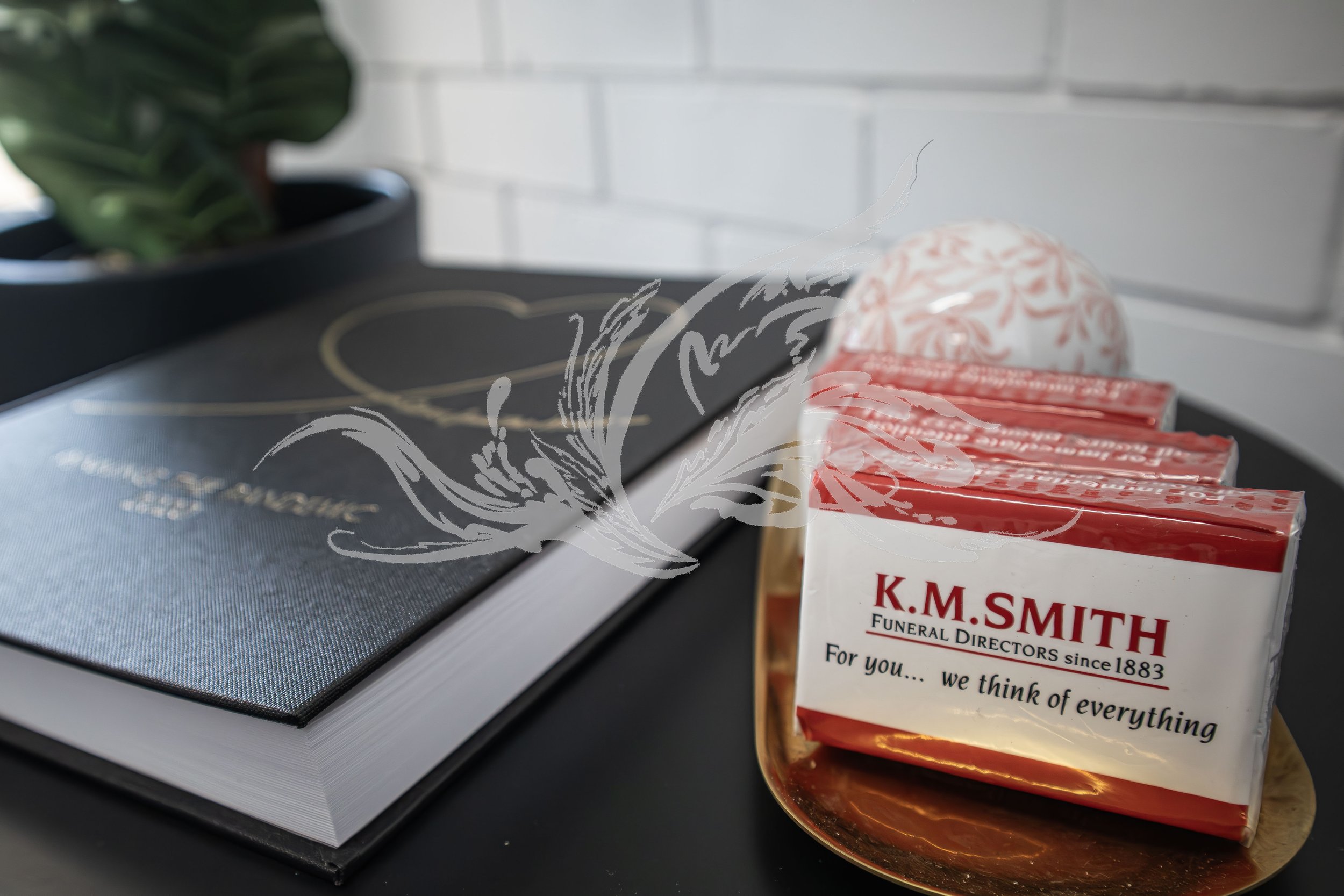 A gift wrapped in clear cellophane with a red and white label from K.M. Smith Funeral Directors, placed on a reflective surface next to a stack of black and white books, with a potted plant in the background and a white brick wall.