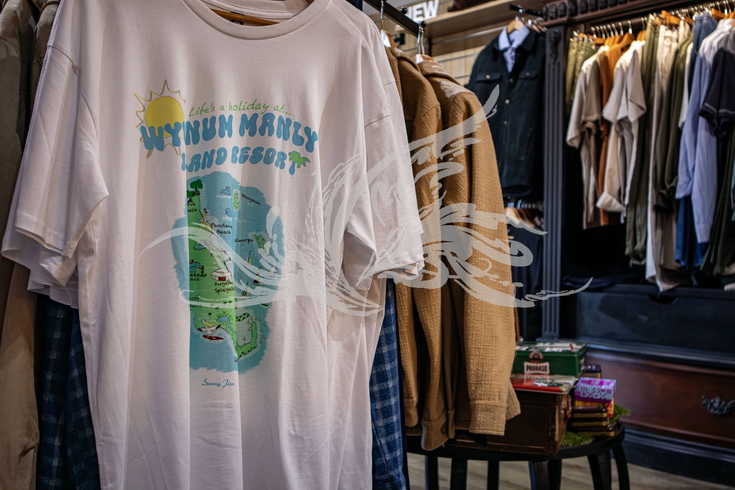 A white T-shirt with a colorful graphic promoting Wynnum Manly and its beaches, hanging on a rack in a clothing store with other shirts and jackets around it.