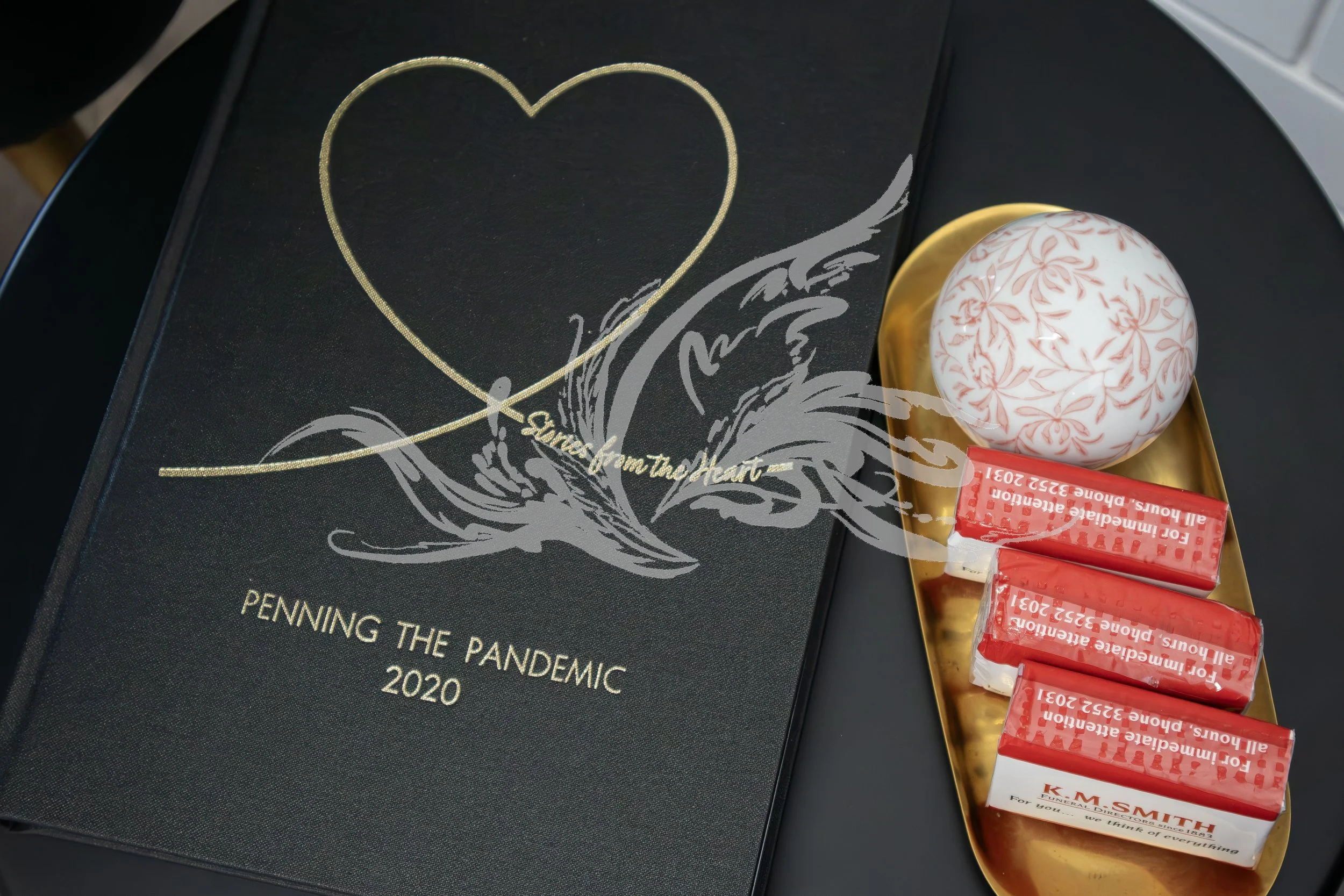 A book titled 'Pennings in the Dark' about the pandemic in 2020, a decorative egg with pink floral patterns, and four red and white Halloween candy bars in a yellow dish.