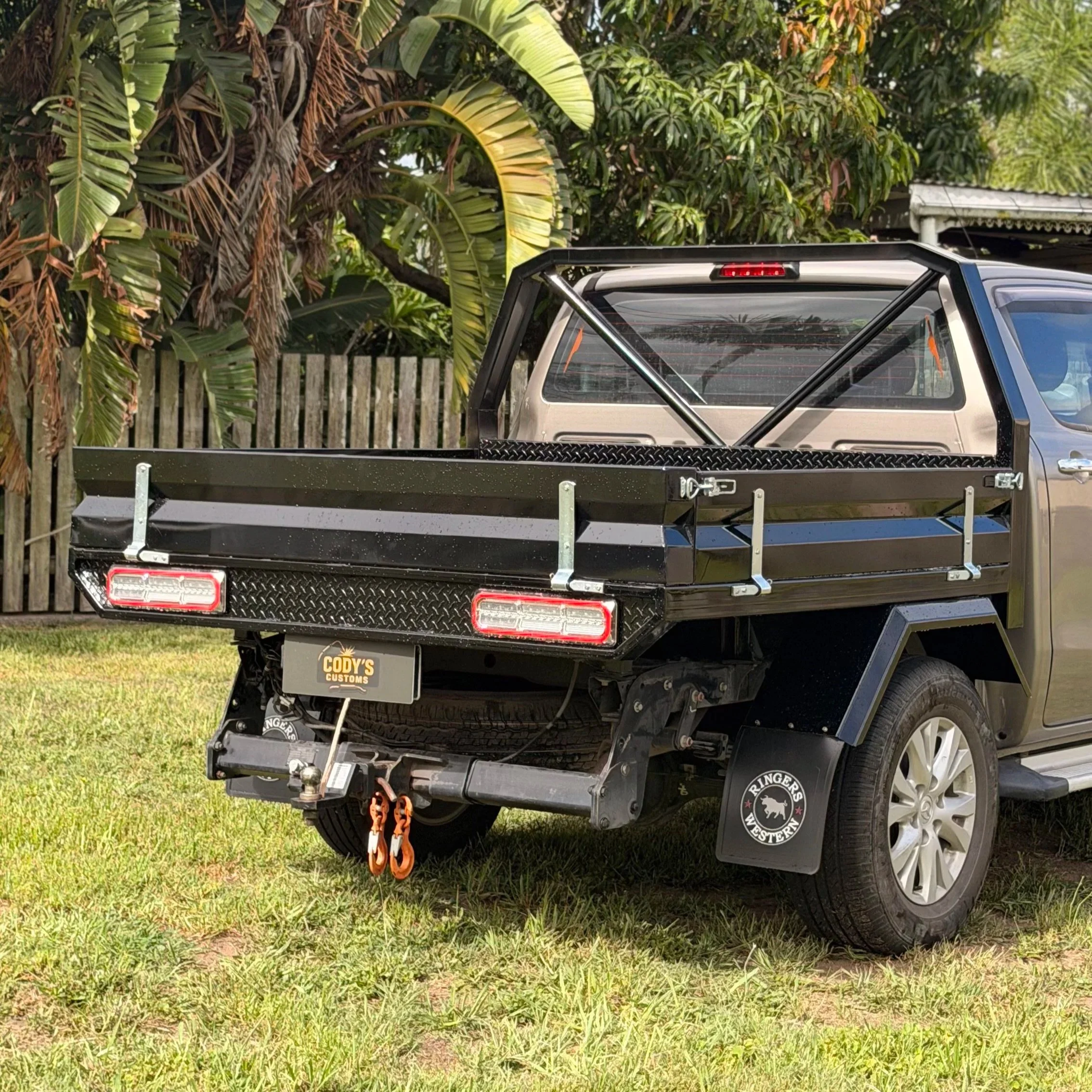 Back of a vehicle with a custom built tray, parked on grass underneath trees and shrubs