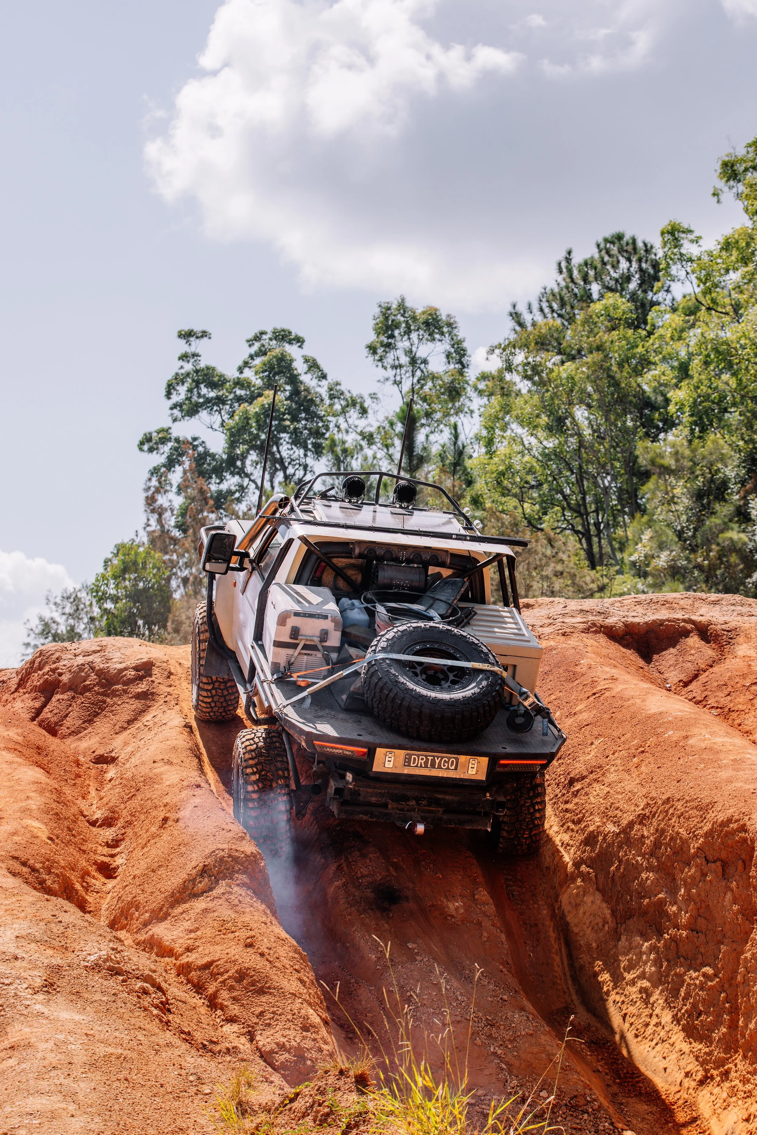An off-road vehicle climbing a steep, rocky dirt hill with tracks in the reddish soil, surrounded by green trees under a partly cloudy sky.