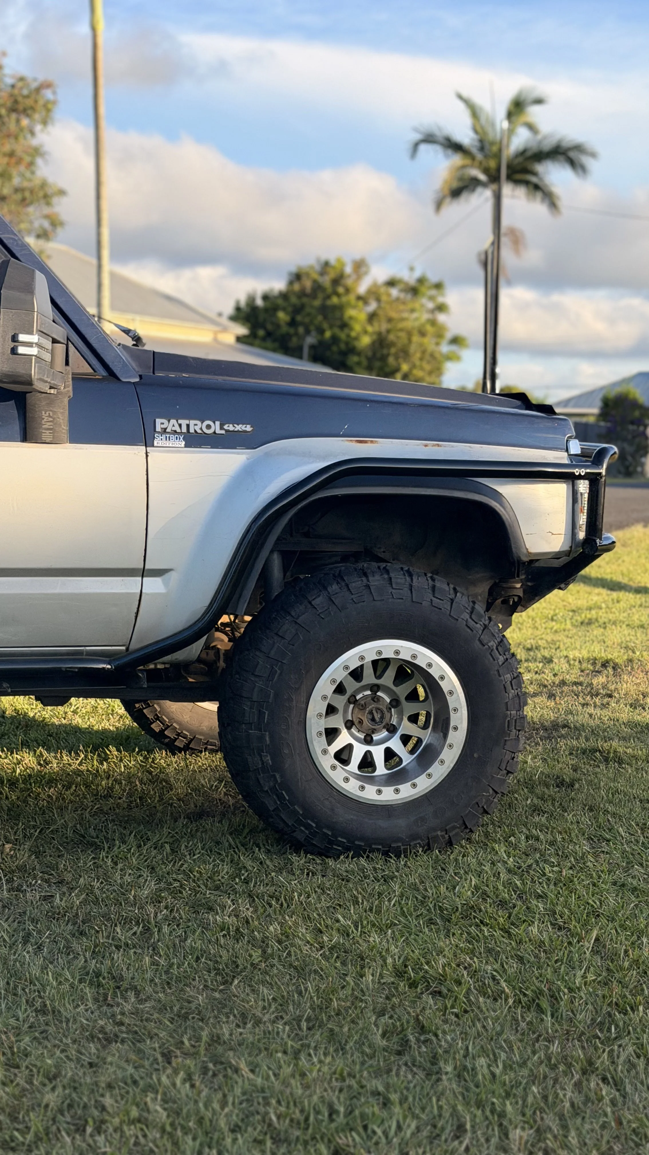 Close-up of a rugged off-road vehicle, showing part of the front wheel, fender, and side panel, with a background of a grassy yard, trees, and a cloudy sky. Custom made scrub bars by Cody's Customs