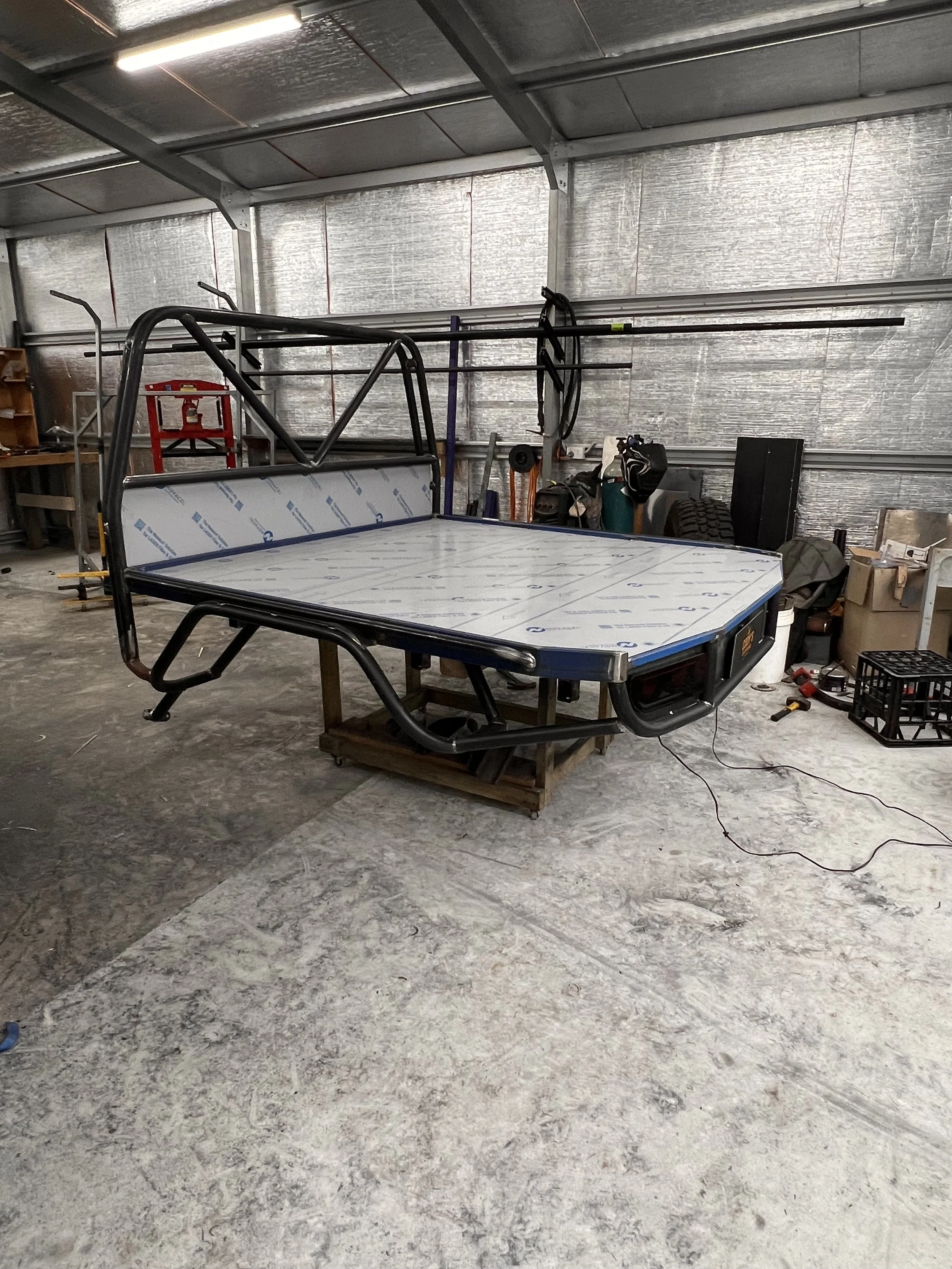 A garage or workshop space with a ping pong table and various tools and equipment stored along the walls. The walls are made of metallic insulation panels, and the floor is concrete.