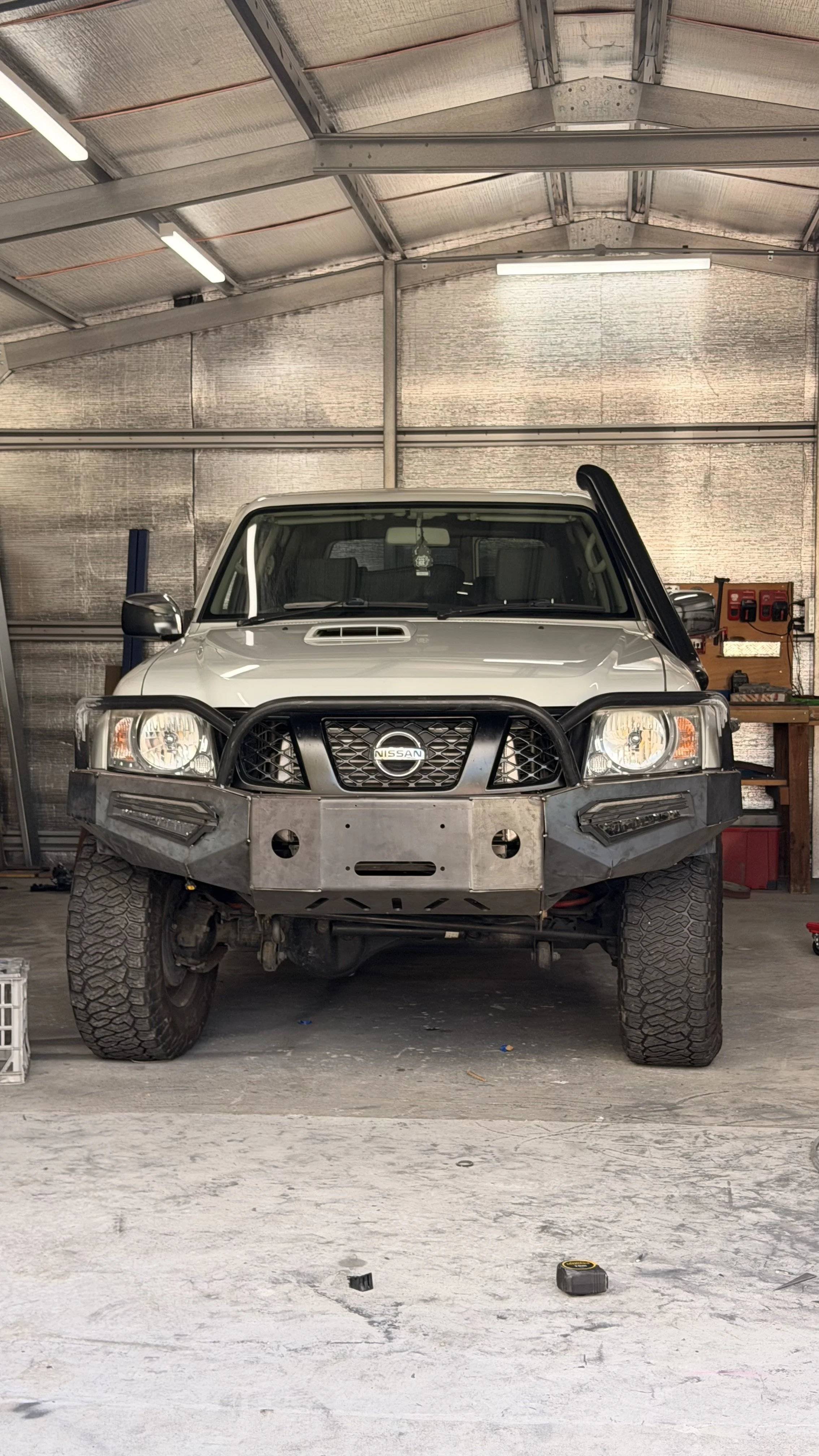 A white Nissan off-road vehicle with a custom built plate bulbar large tires parked inside a garage with a metal ceiling and walls. The garage has tools and equipment on a wooden workbench and a tool chest to the right.