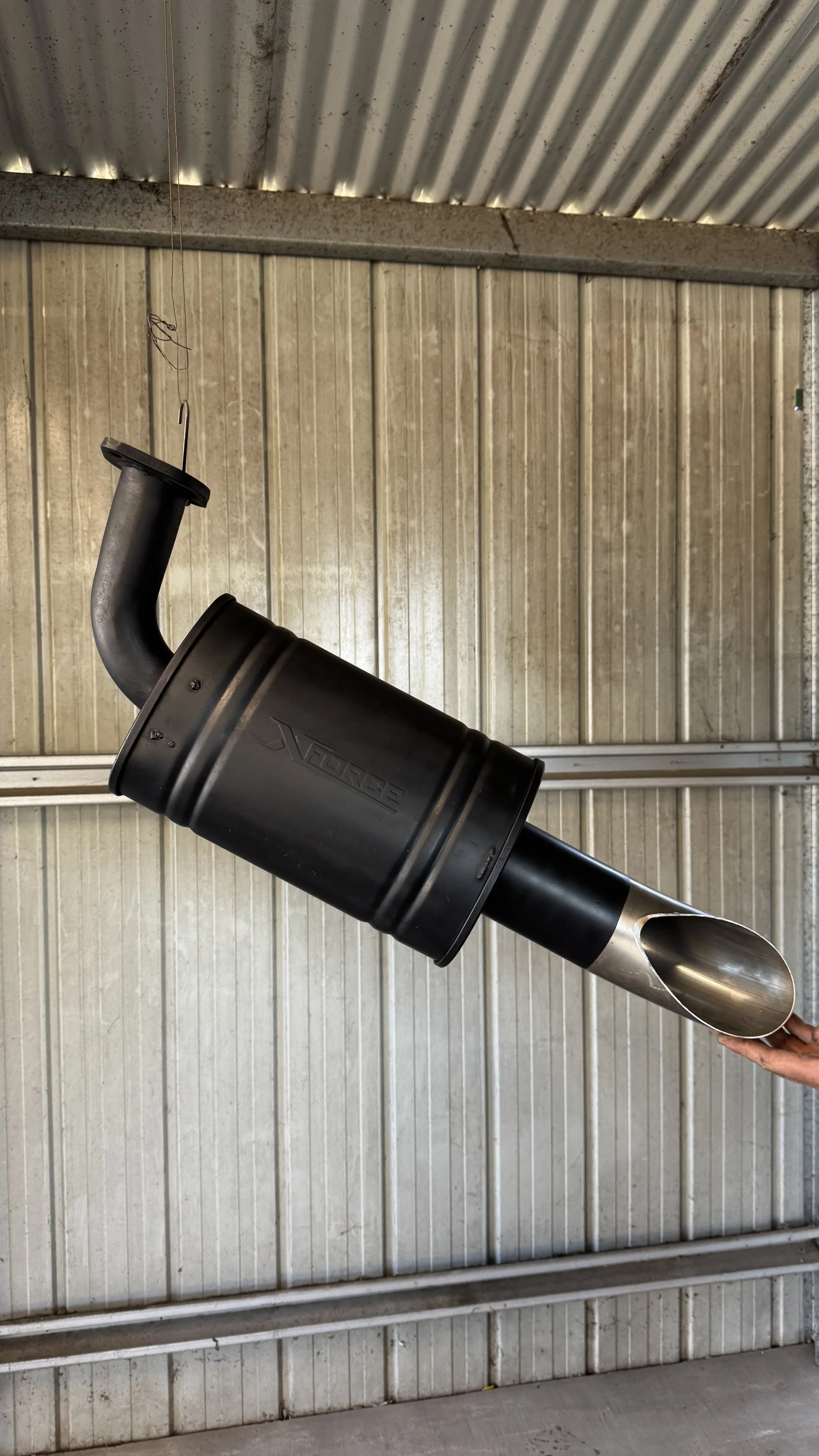 A black aftermarket car exhaust muffler with a curved pipe and a laser-etched brand logo being held in a person's hand against a metal garage wall.