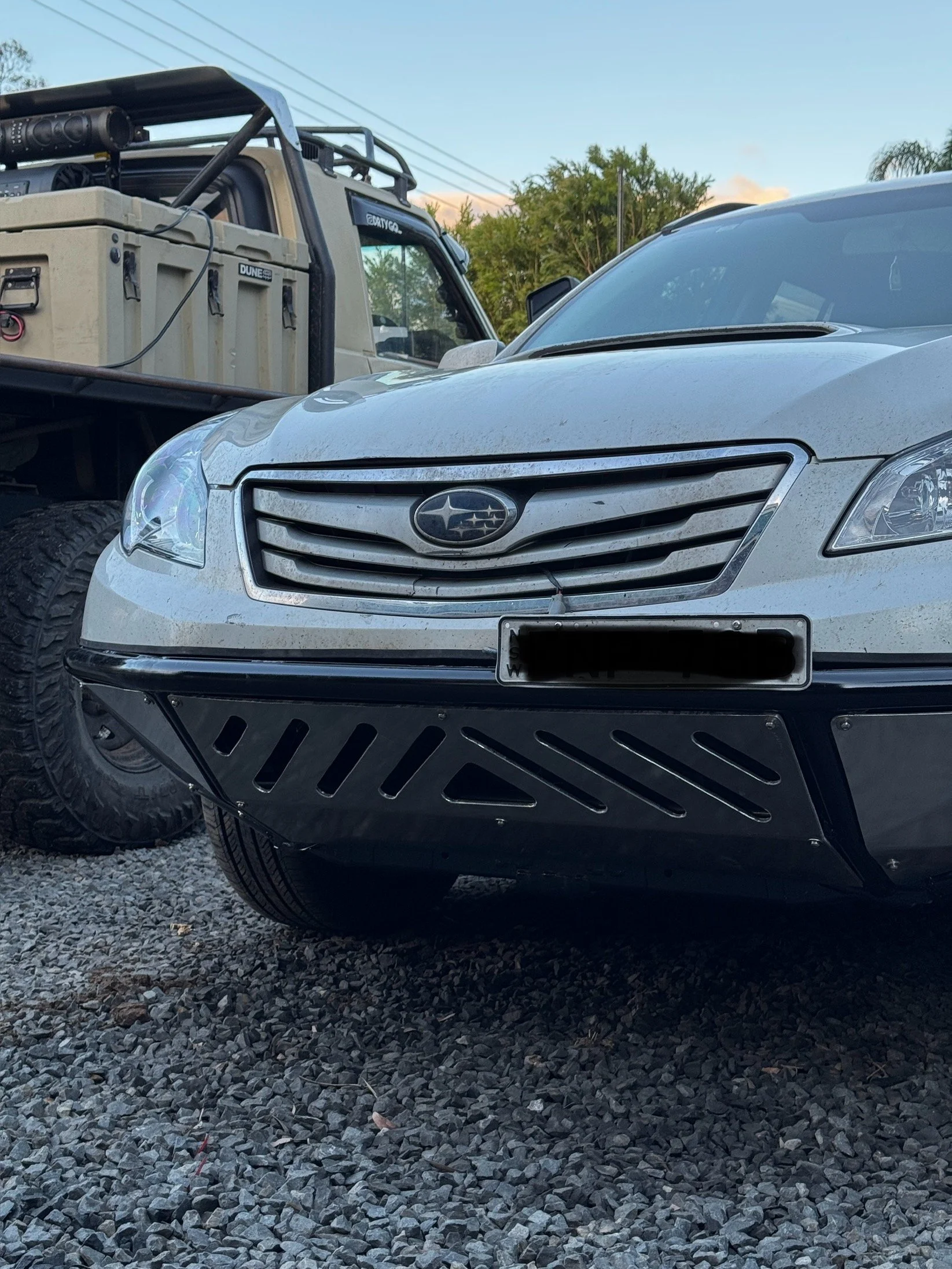 The image shows the front view of a silver Subaru car with a custom made bumper bar parked on a gravel surface, with the grill and headlights visible, and part of another vehicle with a cargo rack and equipment in the background.
