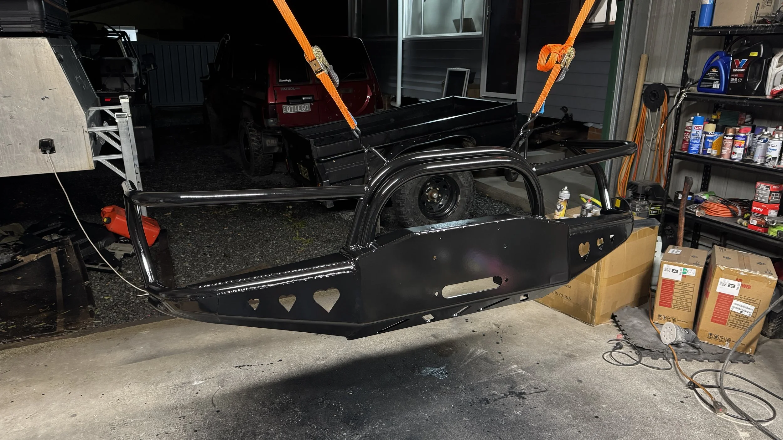 A black custom made bulbar with heart-shaped cutouts being suspended by orange straps in a garage surrounded by tools, automotive supplies, and a vehicle in the background.