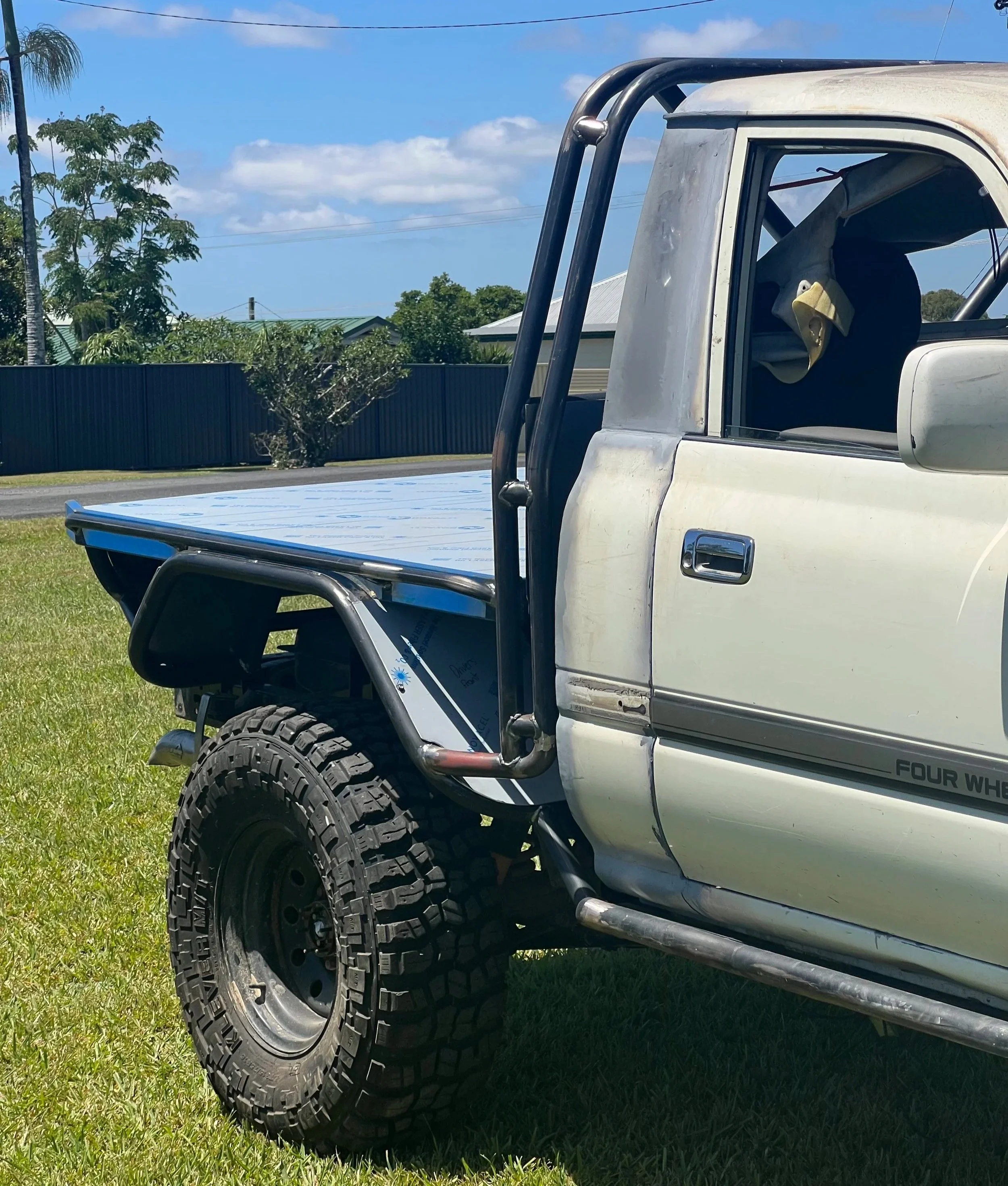 Close-up view of the front left side of a white chopped 80 series parked on grass with large off-road tires, a black roll bar, and a paper partially visible under the tire, with blue skies and trees in the background.