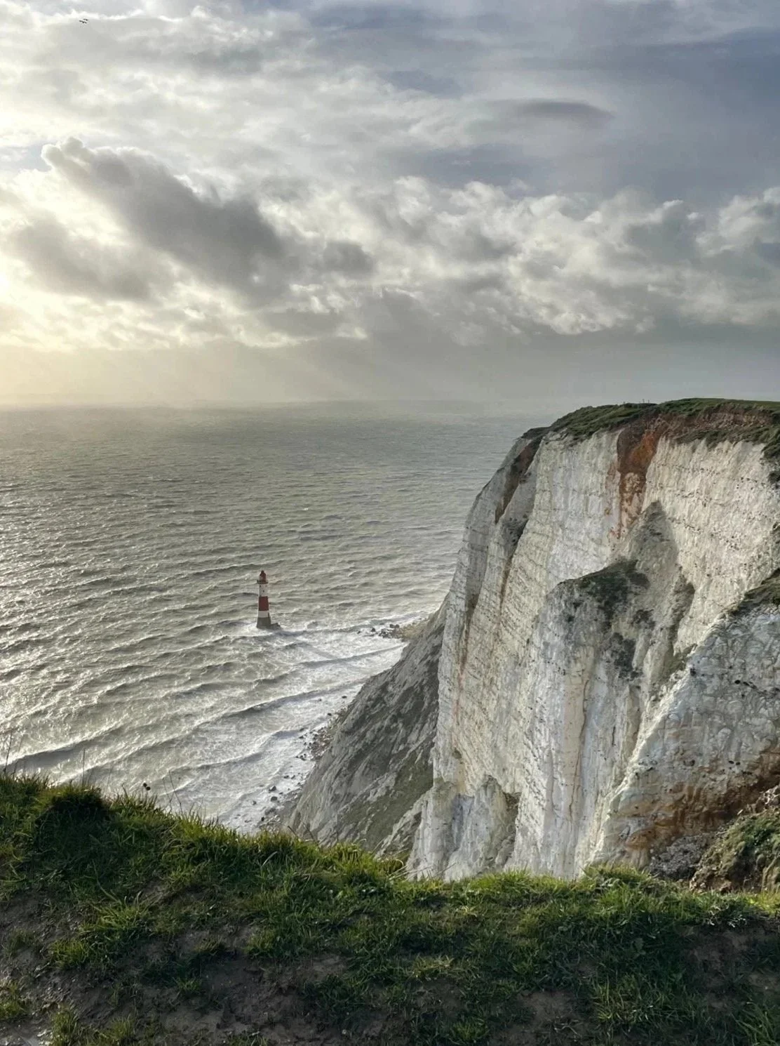 Cliffs overlooking the ocean with a lighthouse in the water and a cloudy sky.