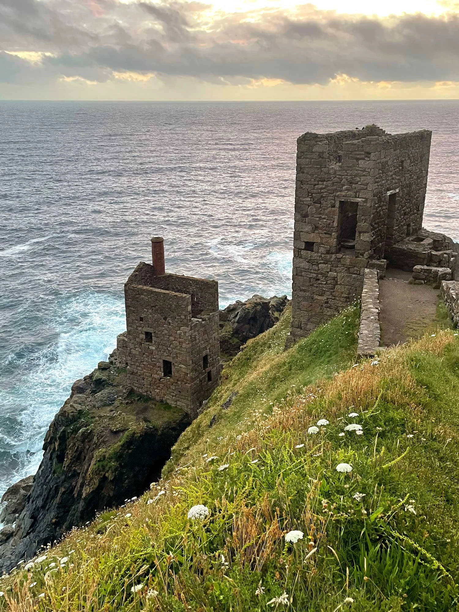 Ruins of an old stone building on a grassy cliffside overlooking the ocean, with a cloudy sky above.