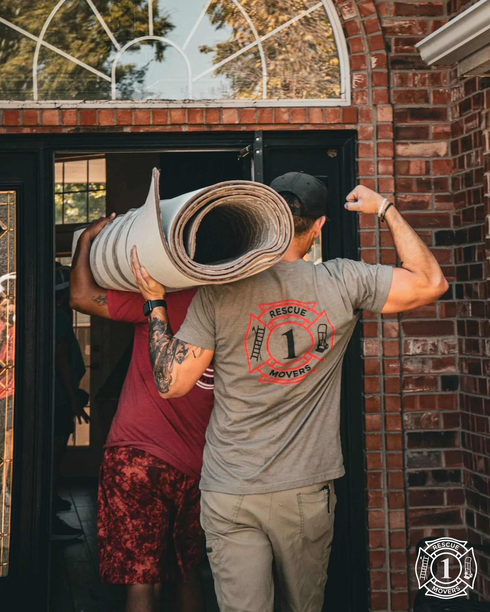 Two people, one with tattoos, moving a rolled-up carpet into a house through a black front door with a brick exterior and a large window above.