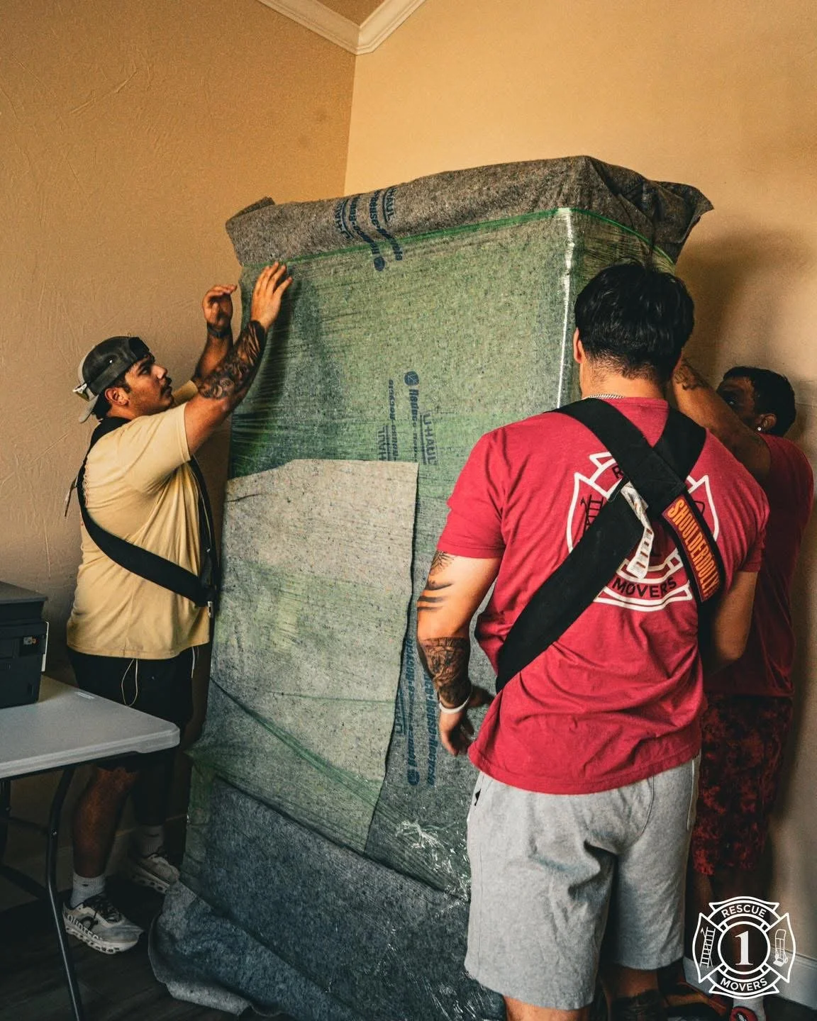 Three men are moving a large, wrapped piece of furniture or object in a room. One man is holding the object, another is looking at it, and a third is adjusting or securing it. The scene appears to be a furniture move or installation.