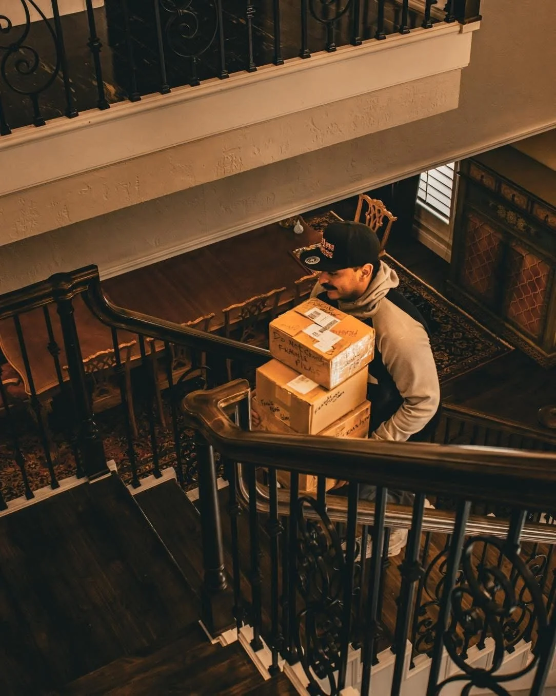 A man standing at the bottom of a staircase, carrying three cardboard boxes, in a home with wooden furniture, a patterned rug, and a sitting area visible.