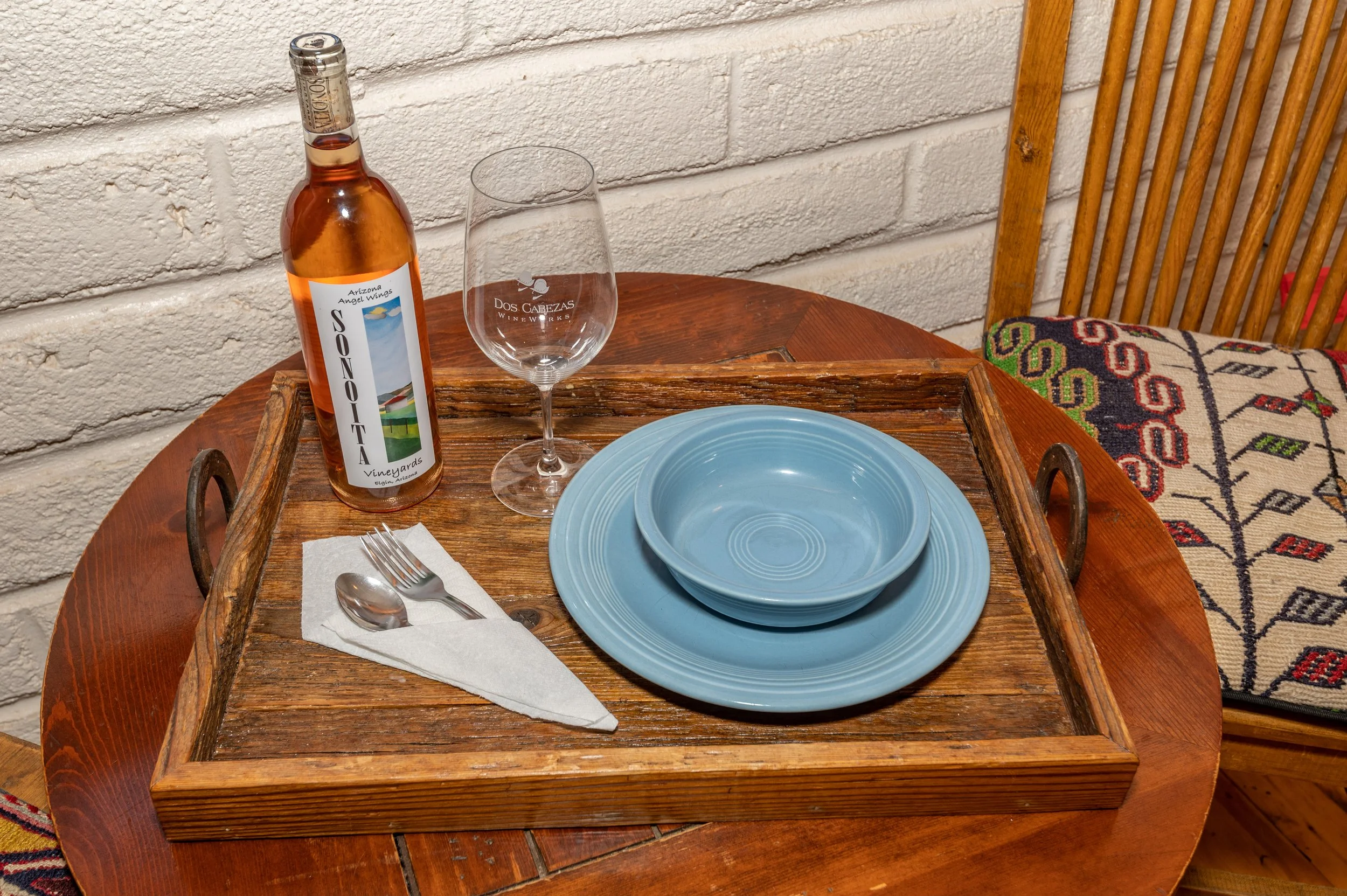 A wooden tray on a round table holds two blue plates stacked, a clear wine glass with logo, a bottle of rosé wine with label 'Sonotal Vineyards', and a fork and spoon wrapped in a napkin. The background includes a white brick wall, a wooden chair wit