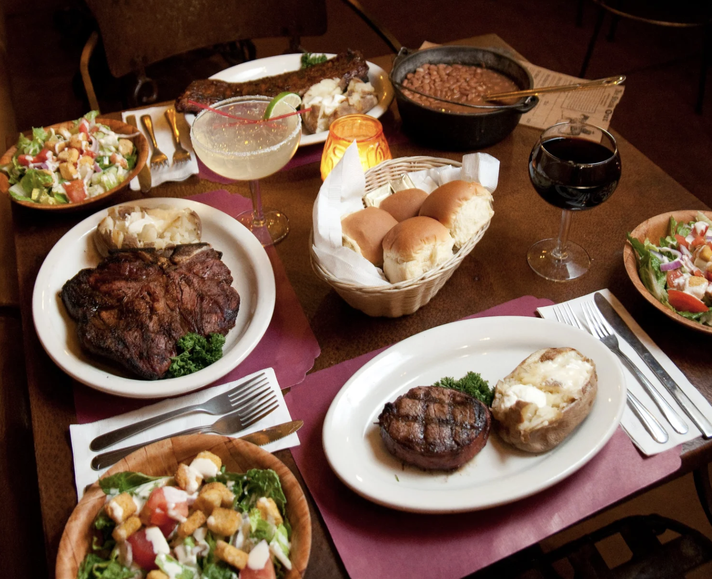 A table with a variety of food including salads, bread rolls, grilled steak, baked potato, beans, and drinks such as red wine and a cocktail.