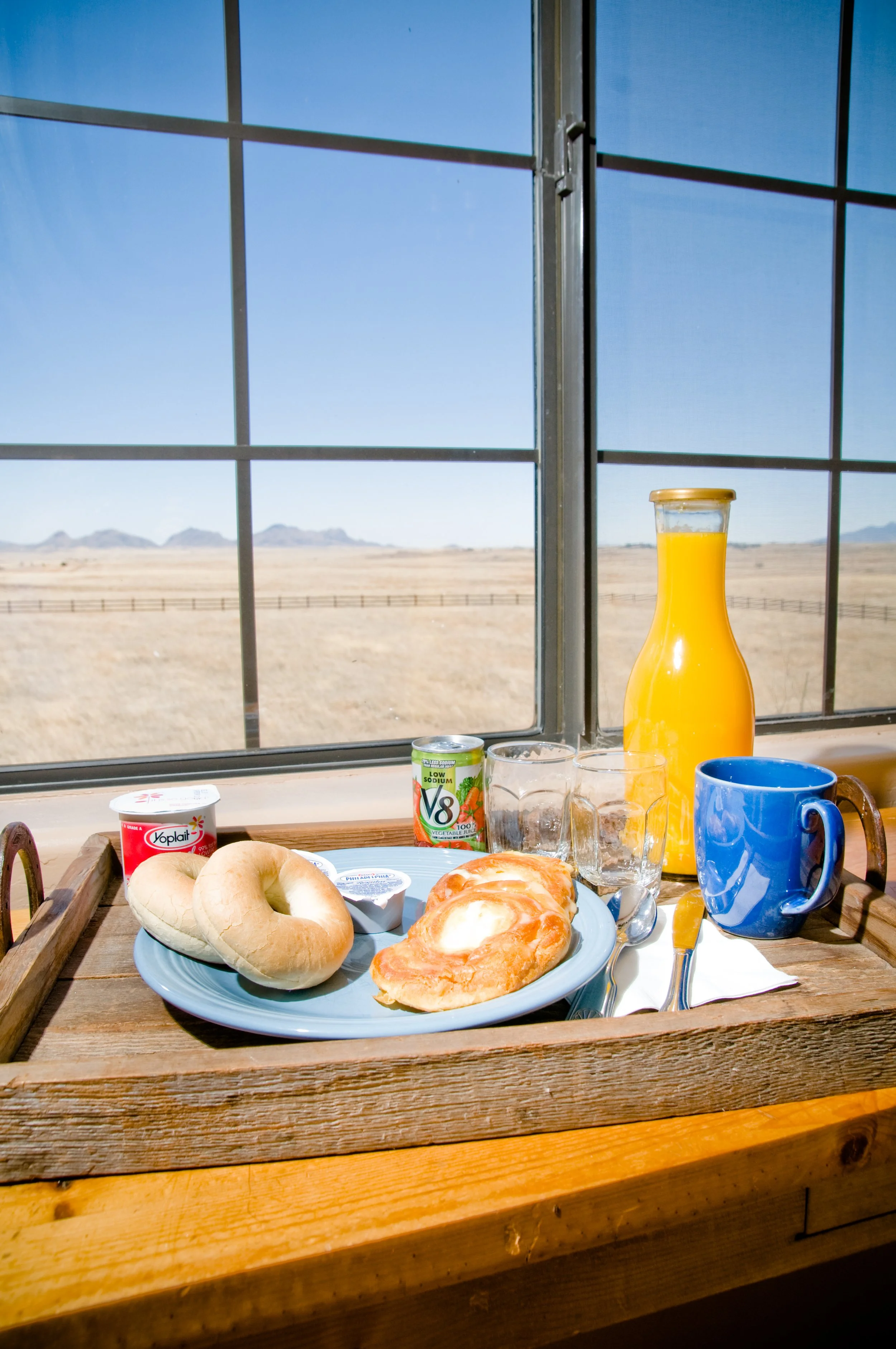 Breakfast tray with bagels, pastries, yogurt, orange juice, and coffee cups, set on a wooden table by a window showing a desert landscape with mountains in the distance.