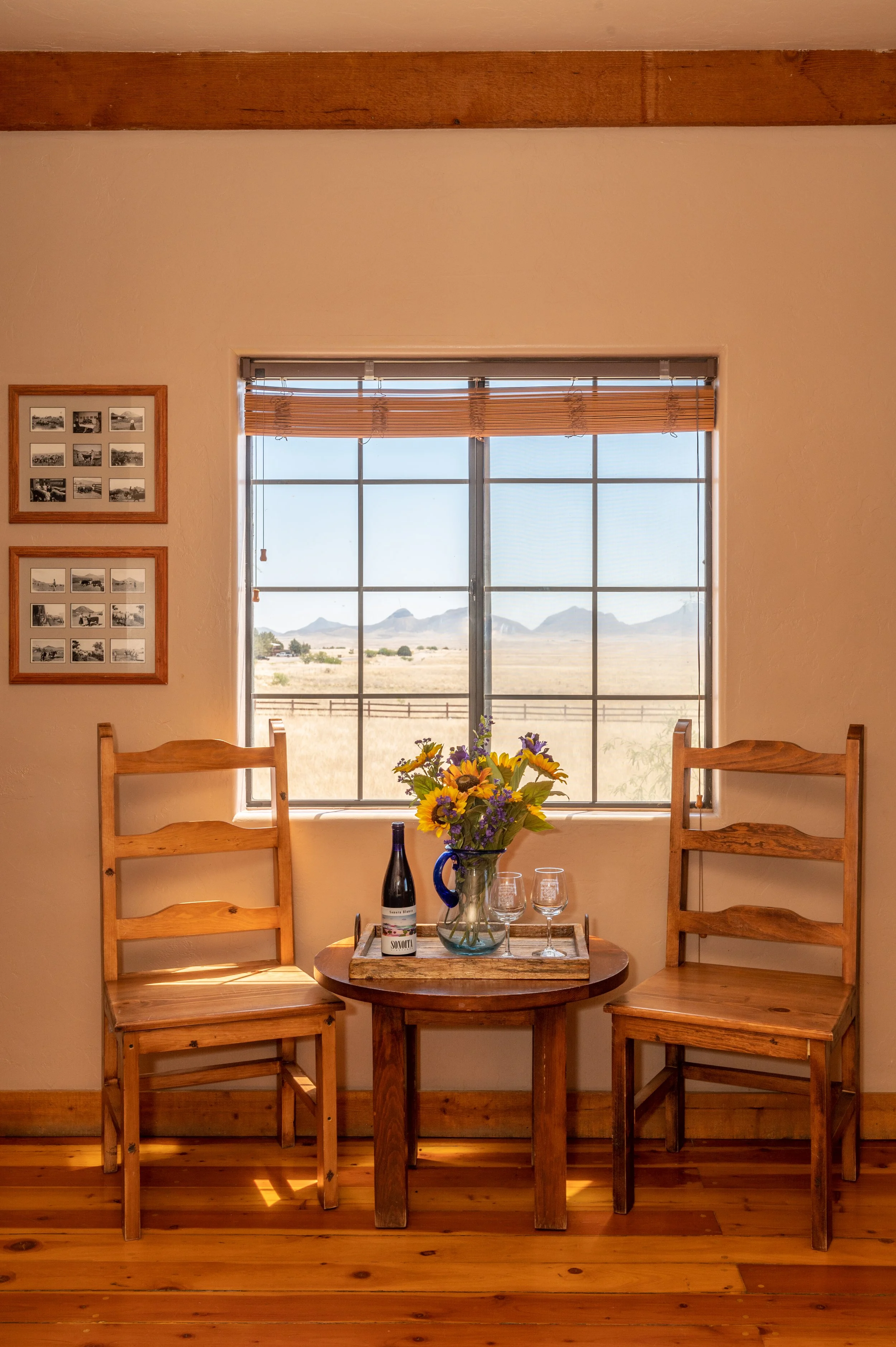 A cozy dining nook with two wooden chairs, a small round table, a vase of sunflowers, a bottle of wine, and two wine glasses. There is a window with a view of a desert landscape and mountains in the background. The wall has framed black-and-white pho