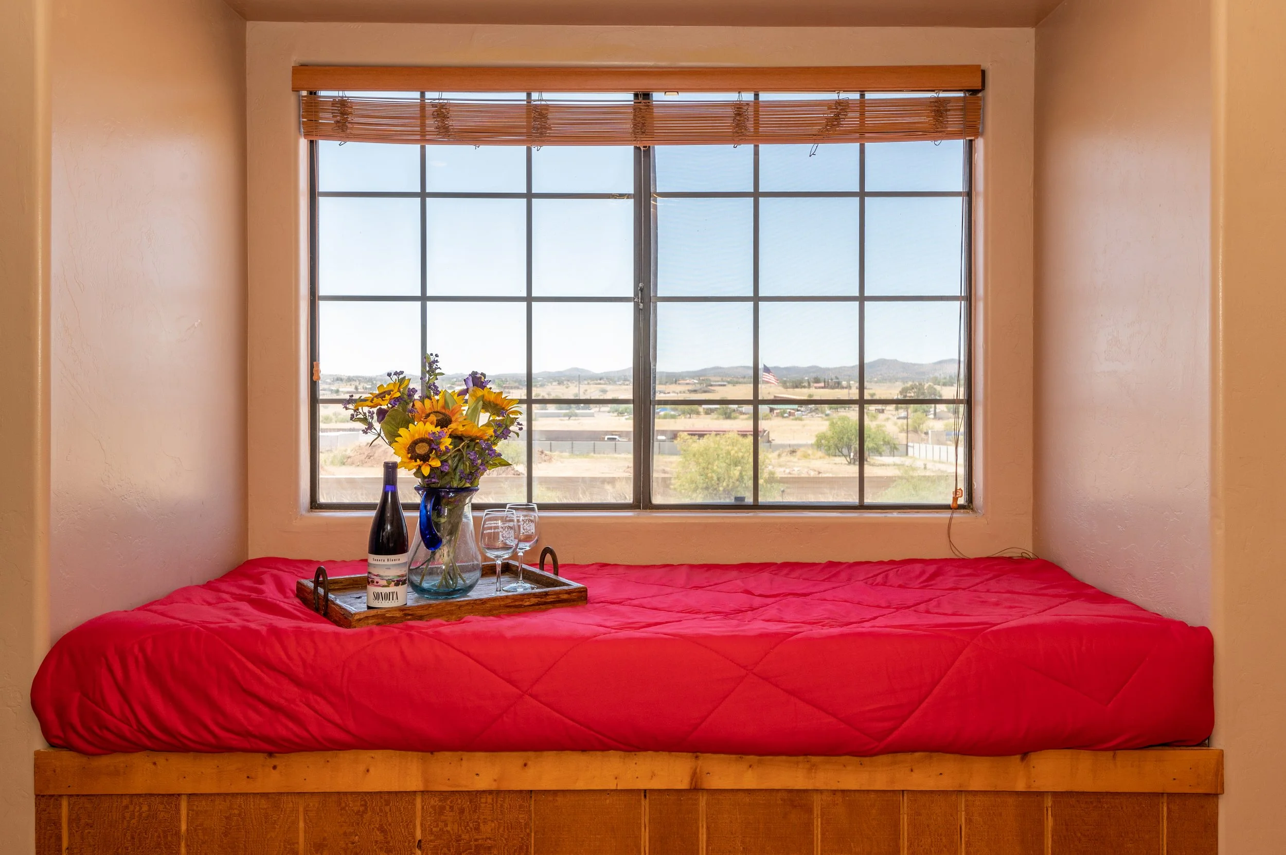 A bedroom with a window view, a bed with a red quilt, a tray with a bottle of wine, two glasses, a vase of flowers, and a small tray on the bed.