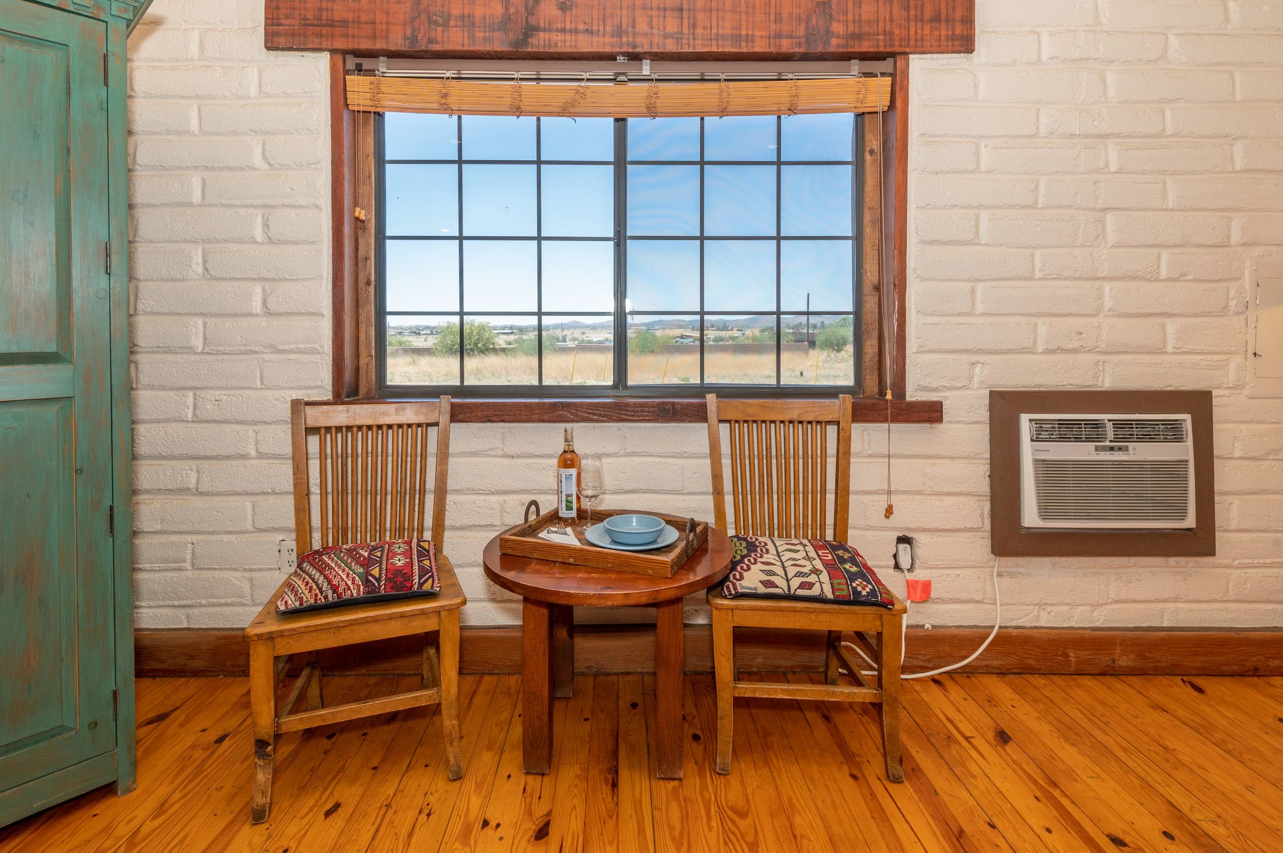 Interior view of a room with a large window, a wooden table set with dishes and two chairs with colorful cushions, a green cabinet, and a heater on the wall.