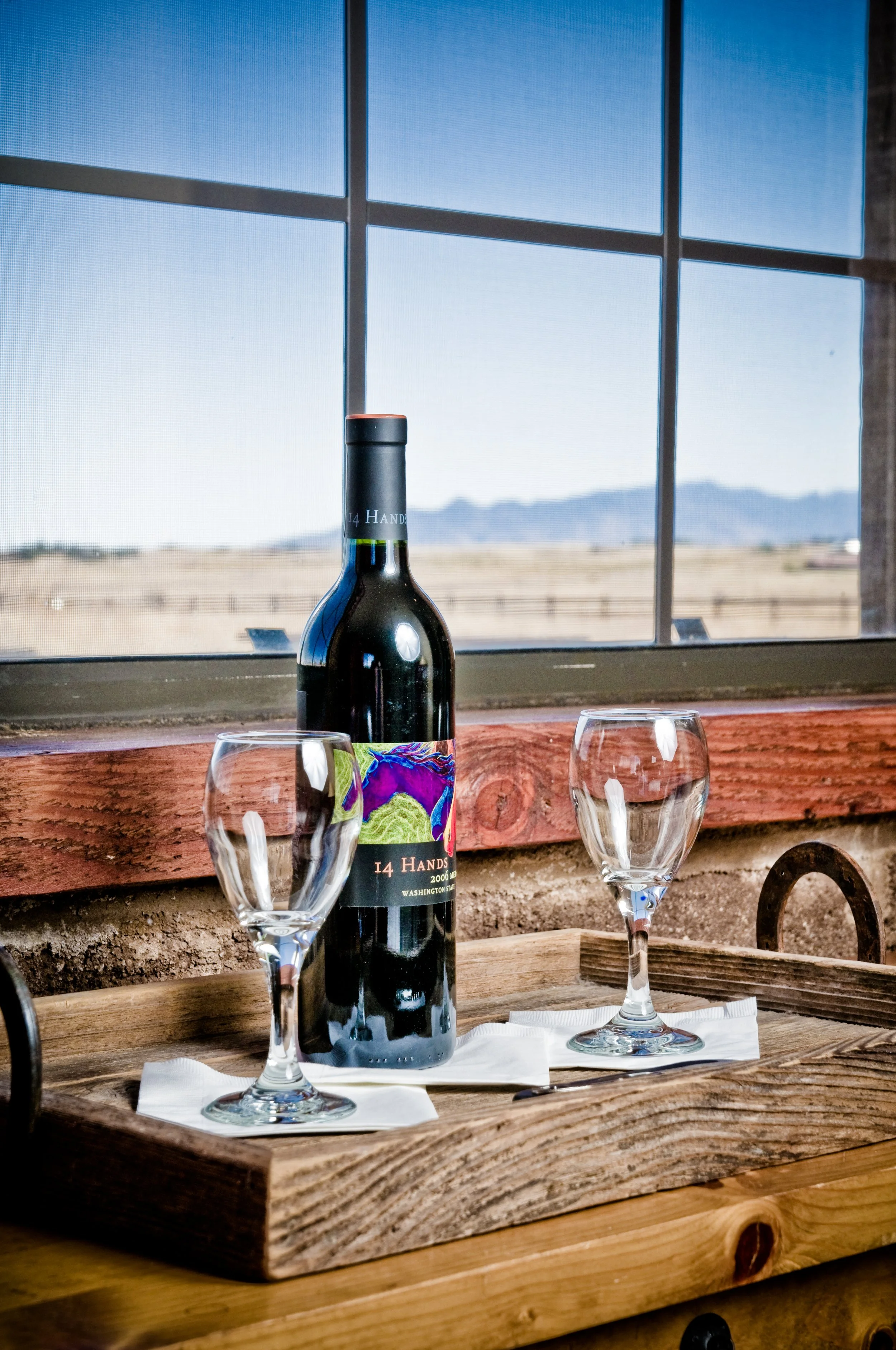A bottle of wine with two empty wine glasses on a wooden tray, set on a table near a window showing a rural landscape with mountains in the distance.