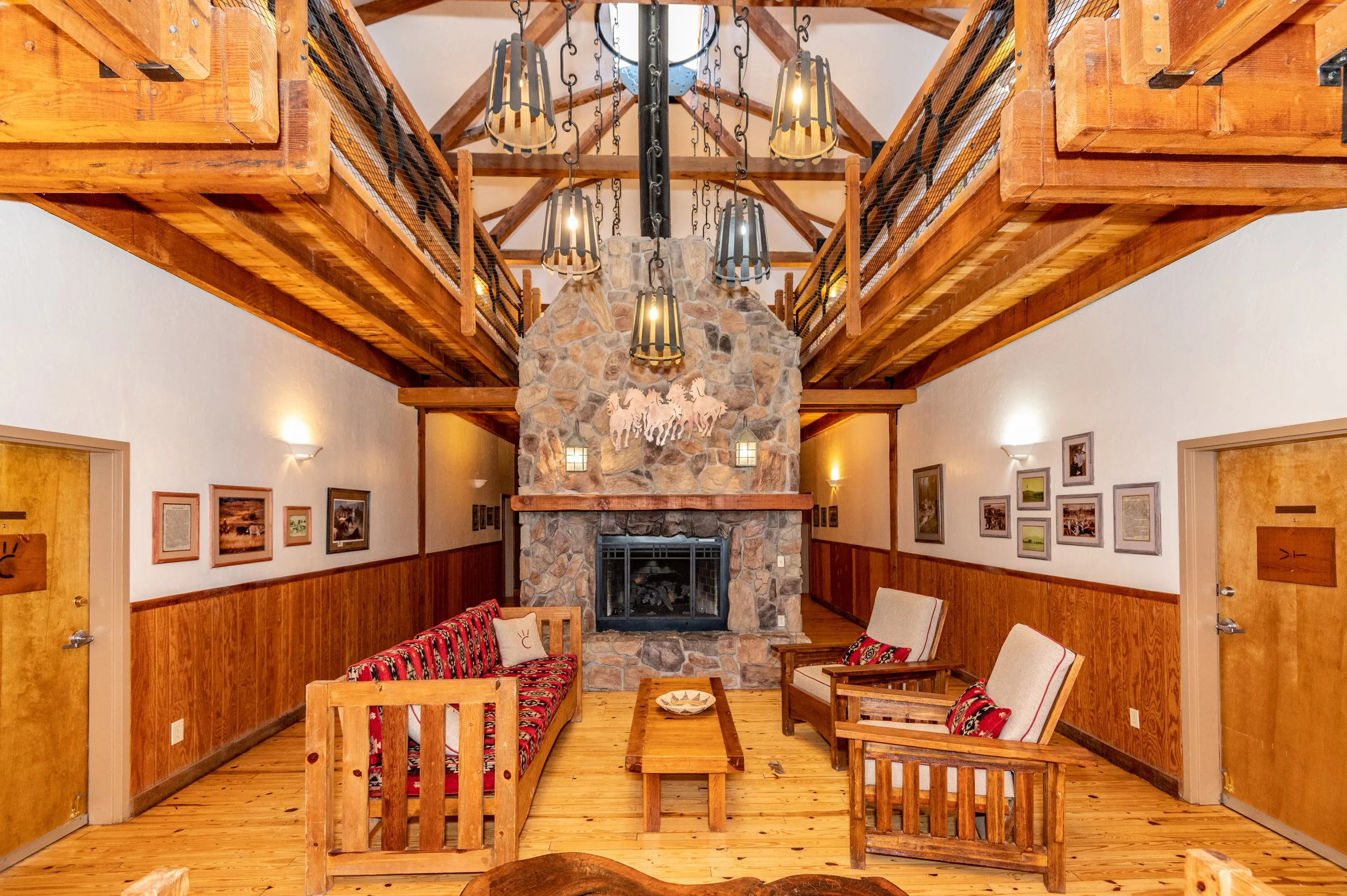 Living room with a stone fireplace, wooden ceiling beams, and rustic furniture, including cushioned chairs and couches with red and patterned fabric, wooden coffee table, and framed pictures on the walls.