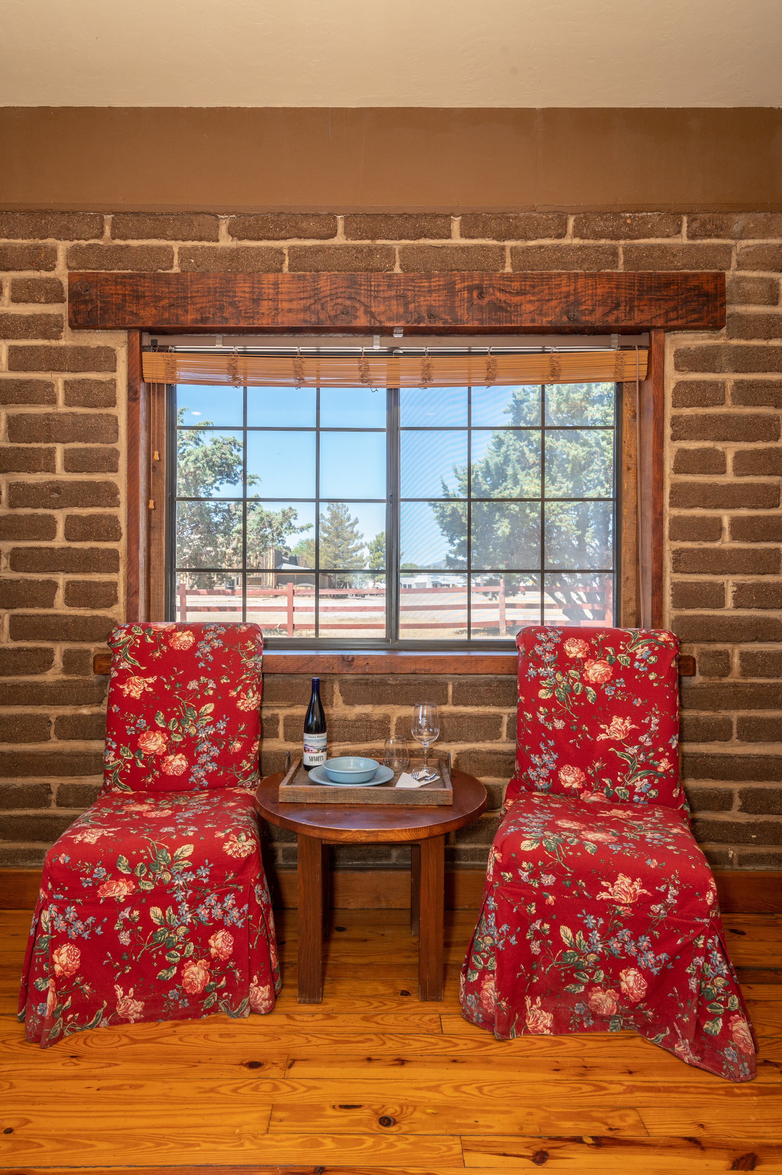 A cozy indoor dining area with two chairs covered in red floral fabric, a small wooden table between them with a tray holding a wine bottle, a wine glass, a bowl, and utensils. Behind the chairs is a large window showing trees and blue sky outside, w