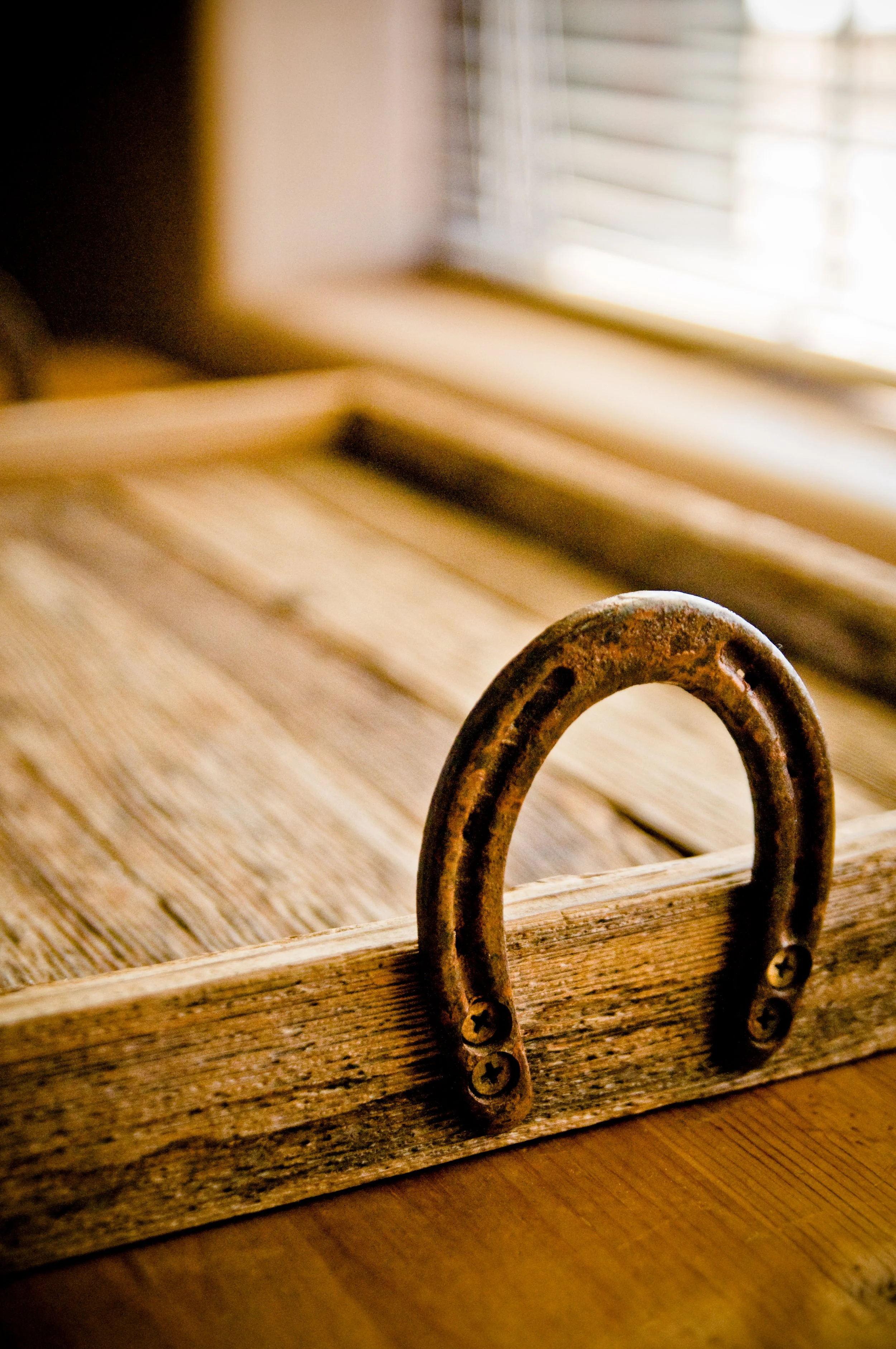 Close-up of a aged, rusty metal hook attached to a wooden surface, with sunlight streaming through window blinds in the background.