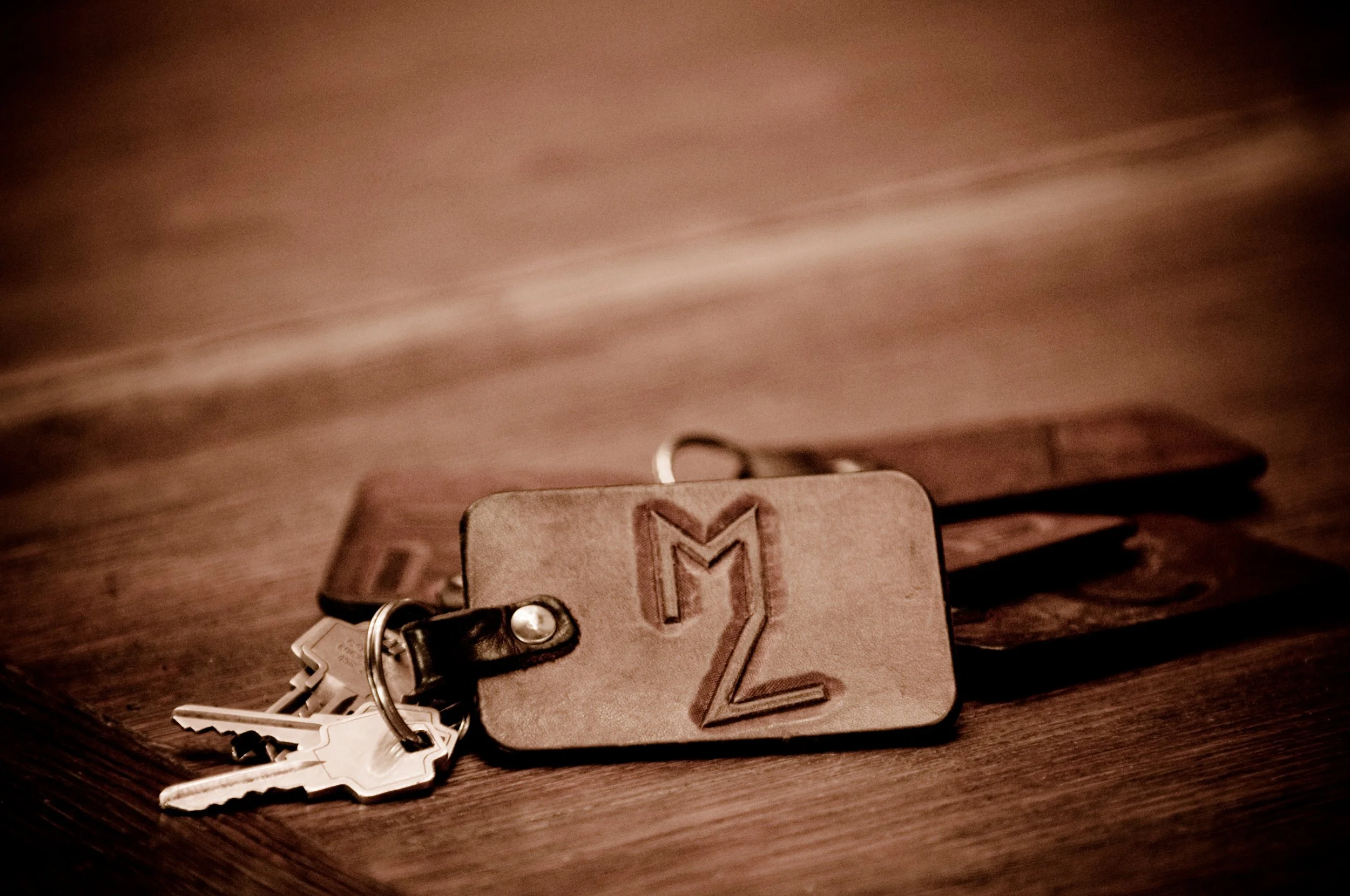 A set of keys attached to a keychain with a beige leather tag that has an embroidered letter M in a dark color, resting on a dark wooden surface.