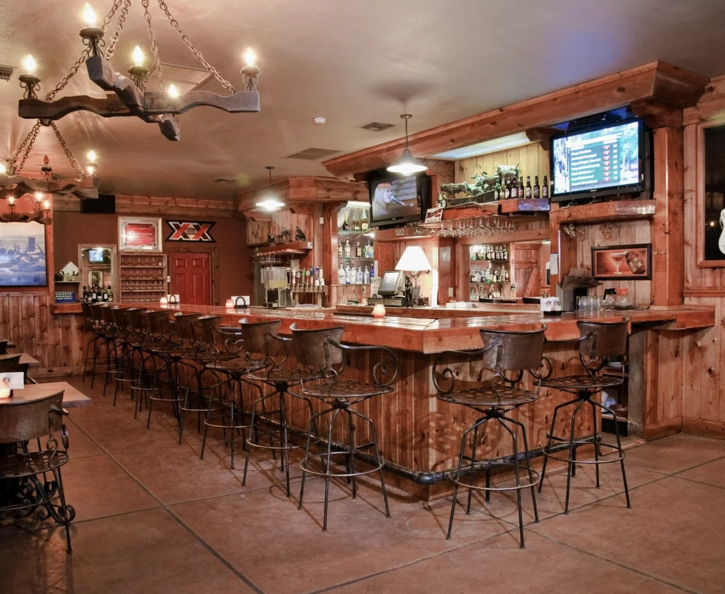 Interior of a rustic bar with wooden decor, high bar stools around a curved wooden counter, televisions mounted on the walls, and chandeliers hanging from the ceiling.