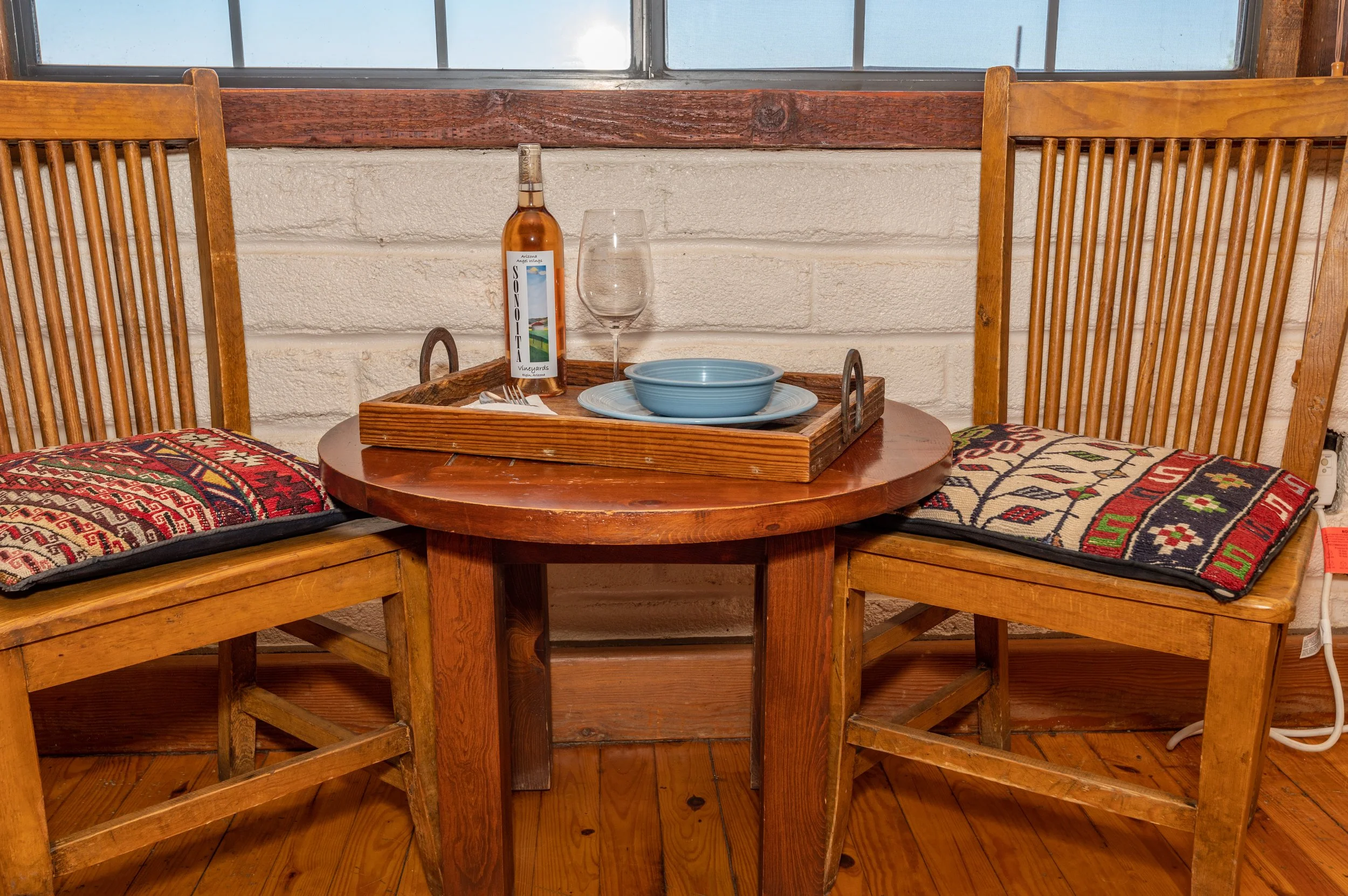 A small round wooden table with two wooden chairs with cushioned seats and patterned fabric, a wooden tray with a bottle of Arizona wine, an empty wine glass, blue bowls, and a set of silverware. There is a window behind the table with a wooden ledge and textured white wall.