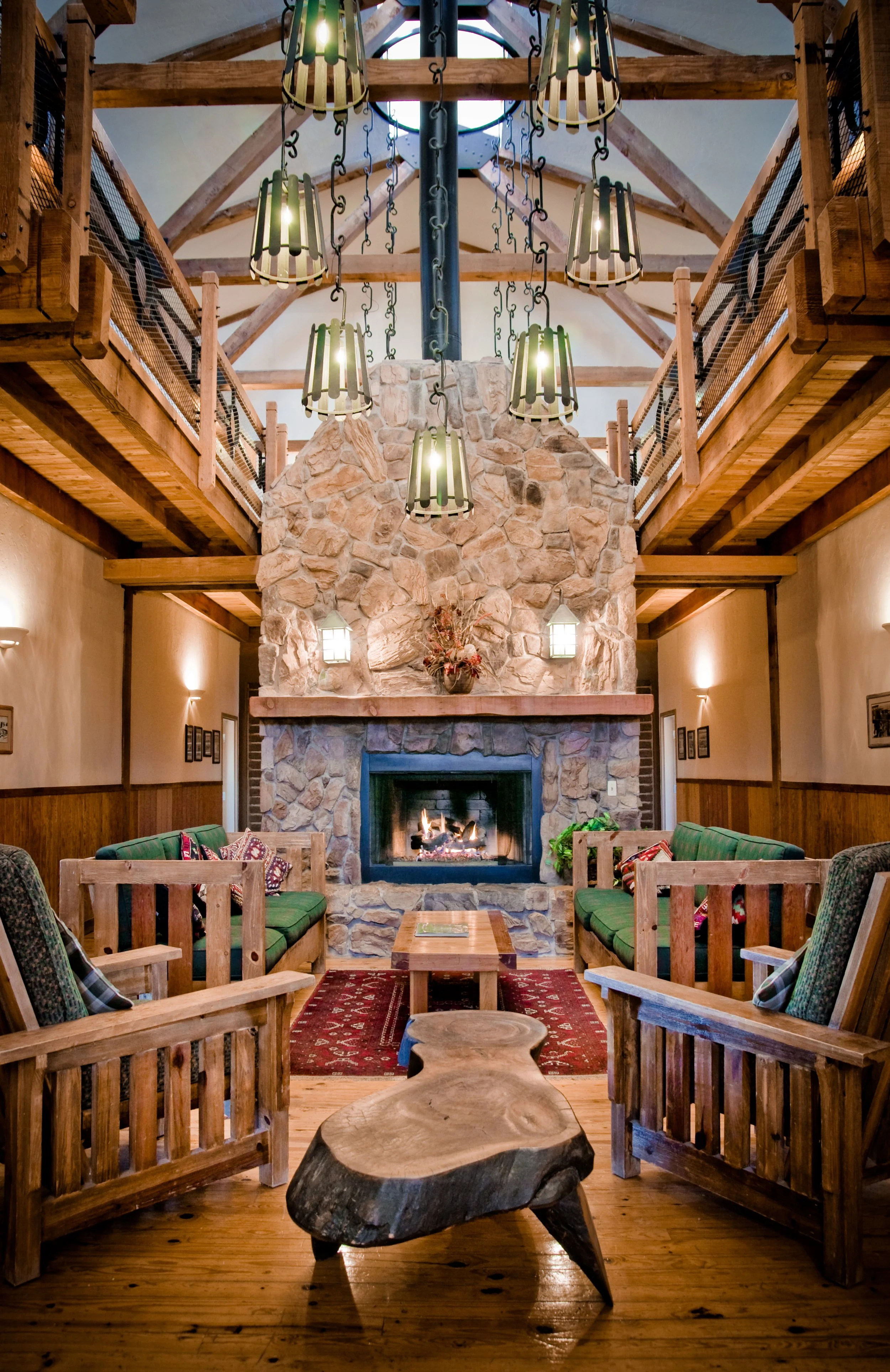 A spacious indoor lodge with wooden beams and a stone fireplace. There are green cushioned chairs arranged around a small wooden table, a red patterned rug, and a large fireplace with a fire burning. Hanging from the ceiling are several black metal chandeliers. There is a decorative wooden table in the foreground and a second-floor balcony with a wooden railing.
