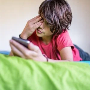 Young boy with brown hair looking at a phone while sitting on a bed with green bedding, wearing a red shirt.