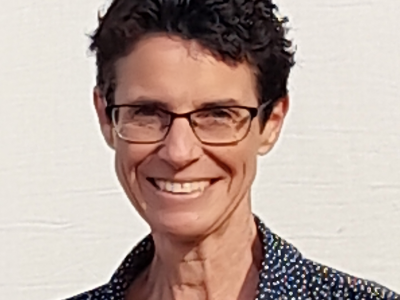 A woman with short dark hair, glasses, and a bright smile, wearing a patterned shirt, standing against a plain light-colored background.