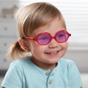 A young girl wearing pink sunglasses and a light blue shirt, smiling in a bright indoor setting.