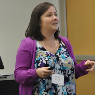 Woman with dark hair wearing a purple cardigan, patterned blouse, and a name tag, speaking in a classroom or conference room.