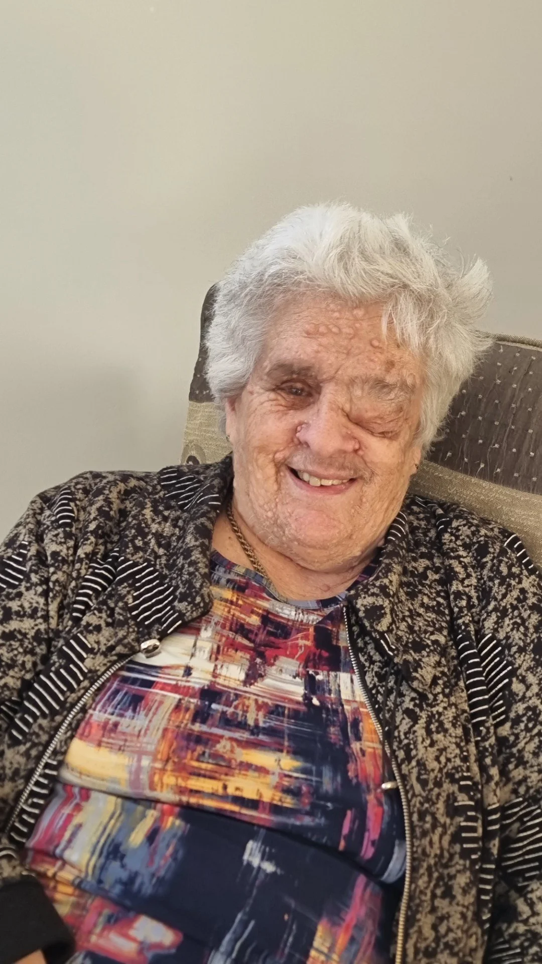 Smiling elderly man with white hair, wearing a patterned jacket over a colorful shirt, sitting on a chair against a beige wall.