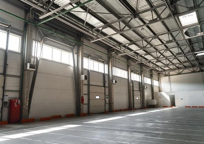 Empty industrial warehouse with large closed garage doors, windows at the top, and a high ceiling with metal beams and roofing.