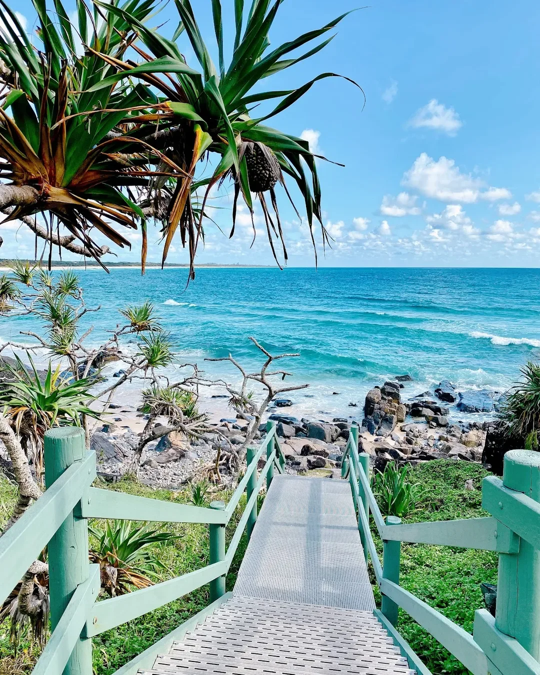 A scenic view of a tropical beach with a wooden ramp leading down to the rocky shoreline, surrounded by lush green plants and trees, with calm blue ocean and partly cloudy sky in the background.