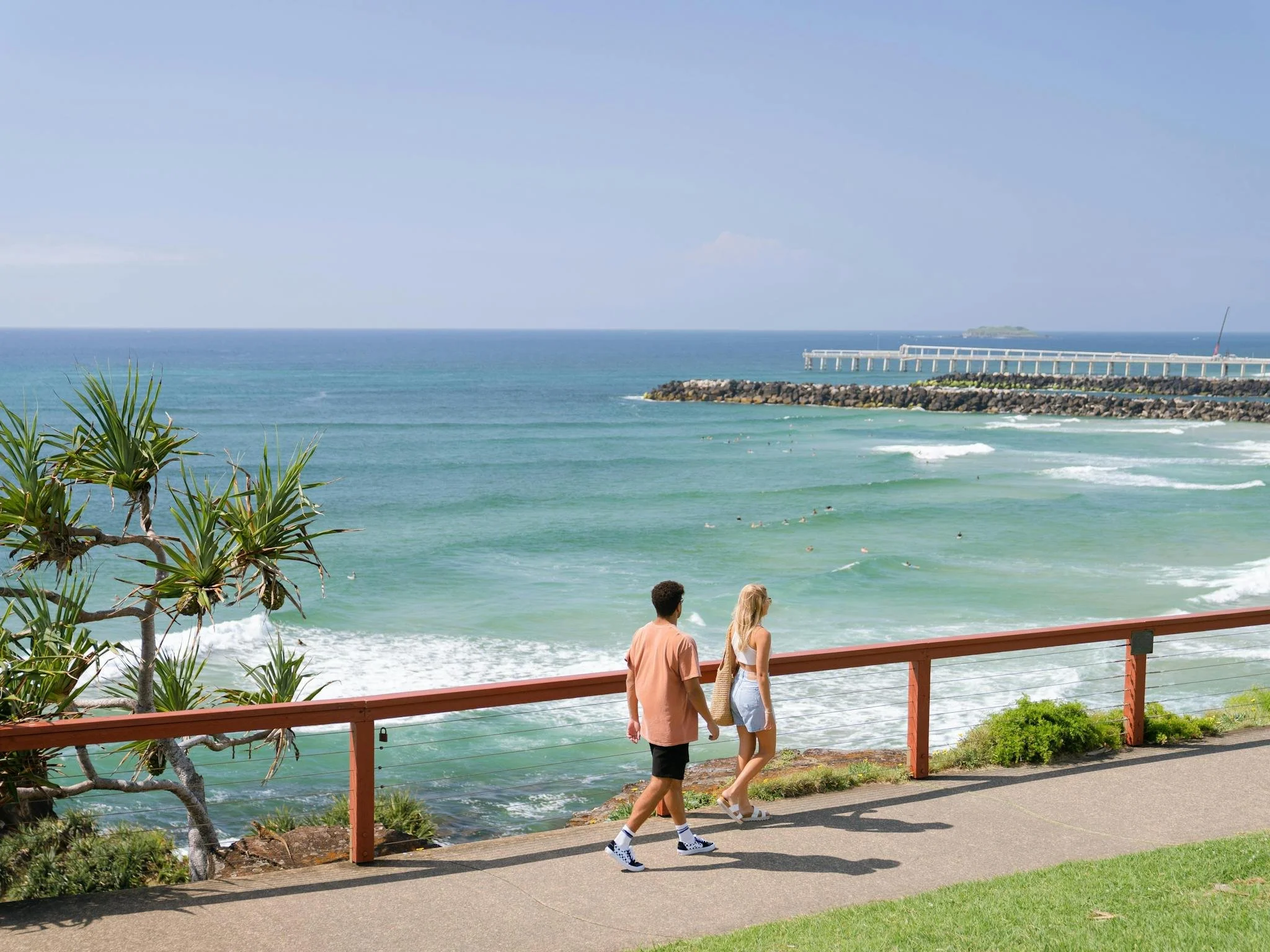 Couple walking along a seaside promenade with a view of the ocean, a pier, and a small island in the background. There are palm trees and green bushes in the foreground.