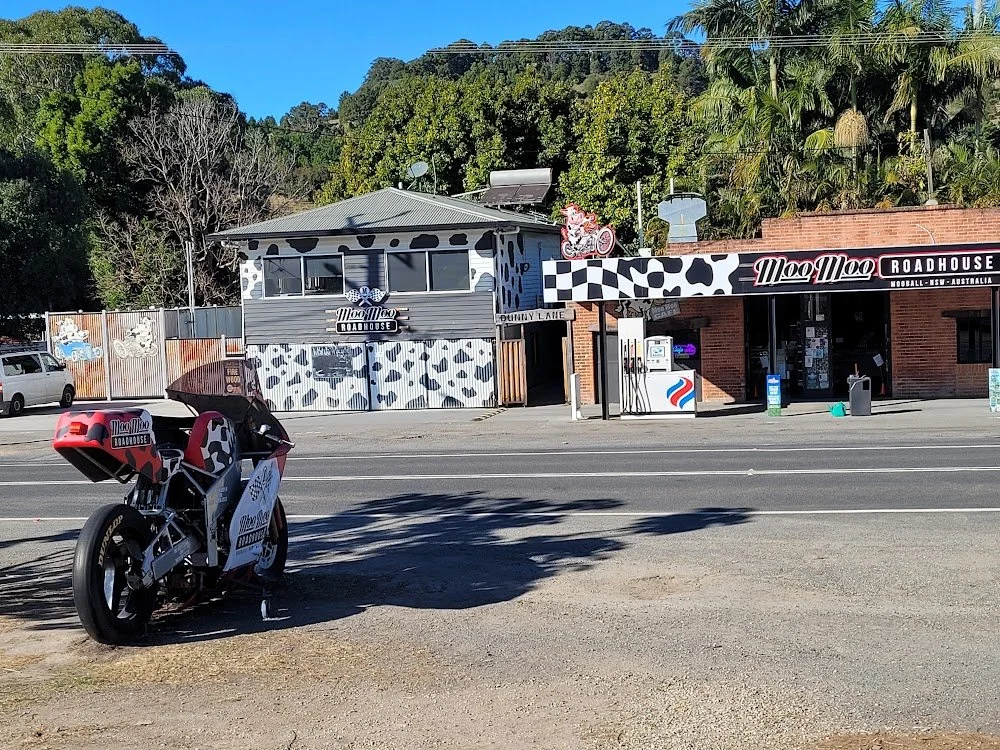 A Mo Mo Roadhouse restaurant featuring a cow-print decor theme, with a motorcycle parked outside on the street and a gas station pump nearby, with trees and a blue sky in the background.