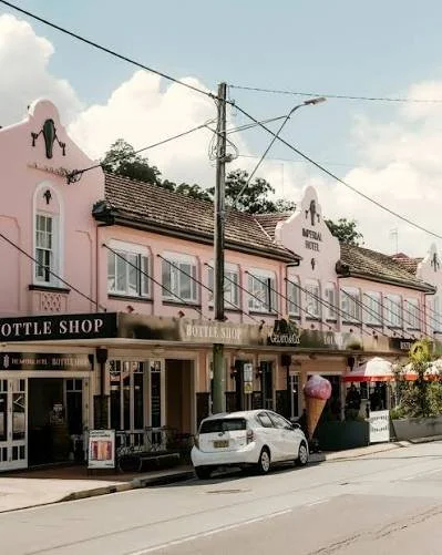 Pink storefront buildings with signs for a bottle shop, a fudge shop, and an ice cream shop. A car is parked in front, and there is a large ice cream cone statue outside.