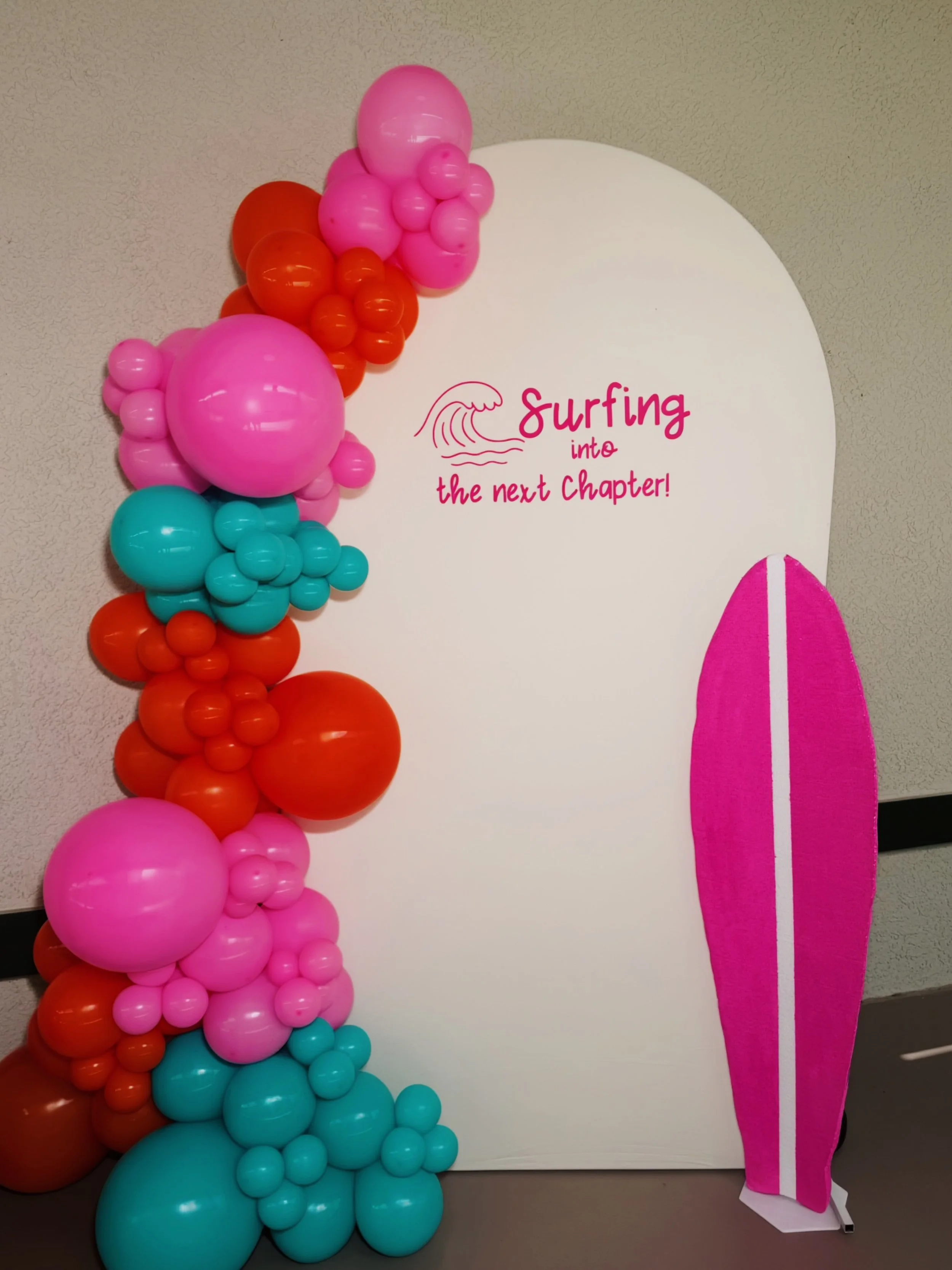 Colorful balloon decoration with various shades of pink, orange, blue, and purple arranged in a cascading fashion next to a white surfboard-shaped sign that reads "Surfing into the next Chapter!" in pink and red lettering, with a wave illustration above the text.