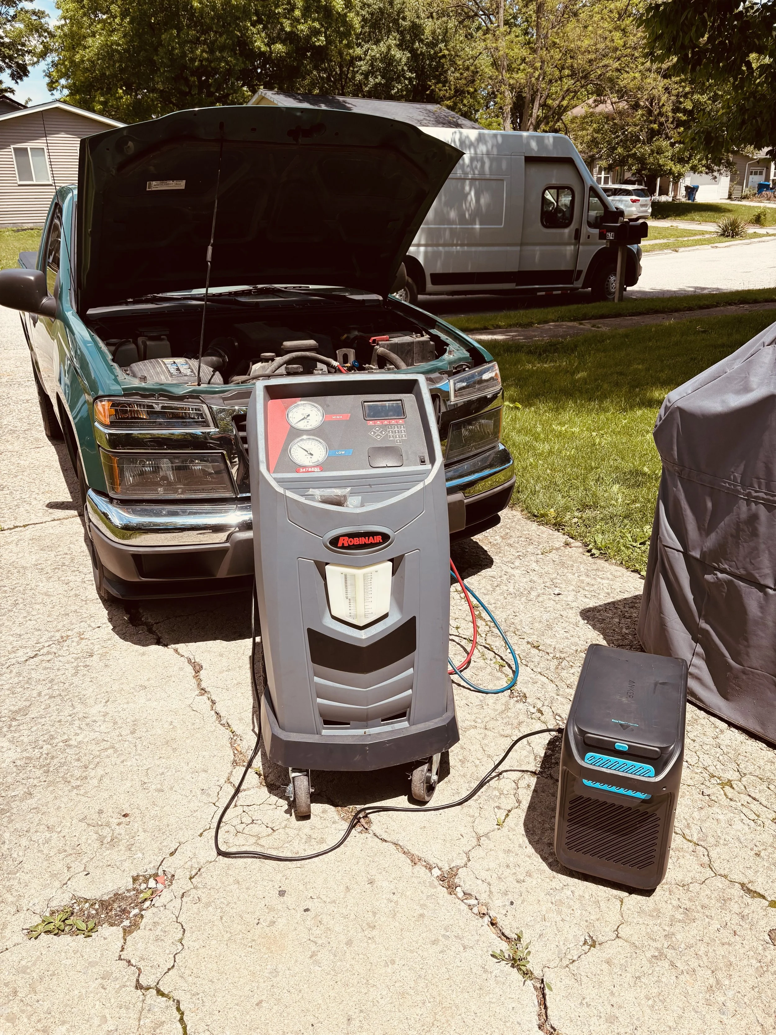 A car with its hood open connected to an AC machine, while the Lorenz Auto Care van is in the background at the customers house